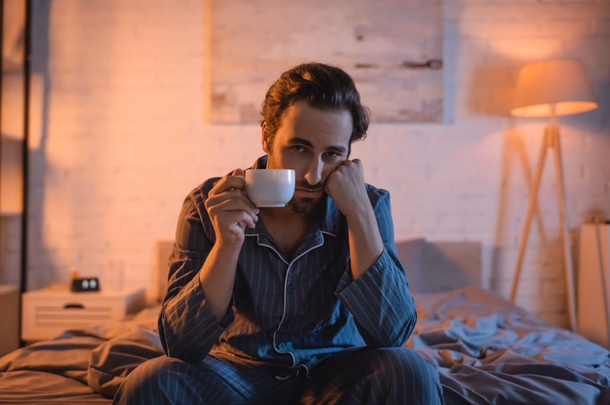 坐在床上睡眠不足的男人很不高兴拿着杯子看着镜头生活方式