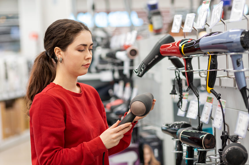 Caucasian young woman with long hair, holding a hair dryer. In the background, shelves with household appliances. The concept of buying household appliances for beauty and self-care.女子手拿吹风机生活方式