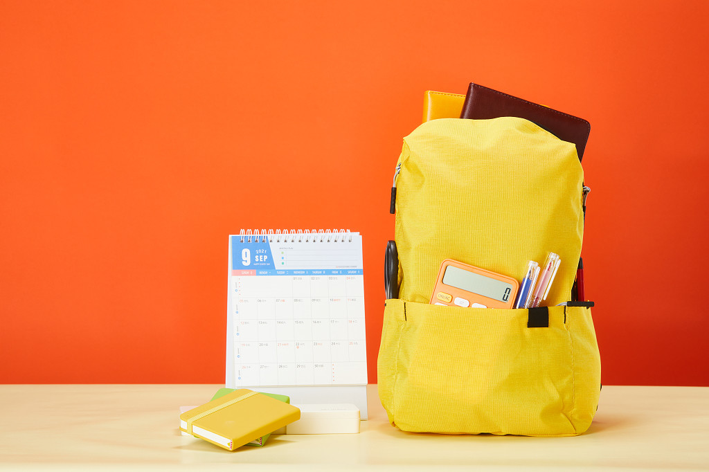开学季橙色背景前装满文具的黄色书包台历学习用品场景配图
