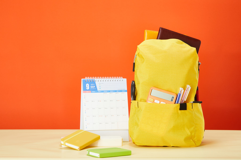 开学季橙色背景前装满文具的黄色书包台历笔记本场景配图
