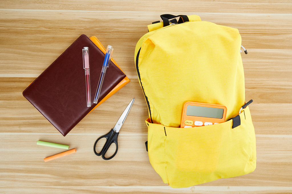 开学季装着计算器的黄色书包文具在木制课桌上的场景配图
