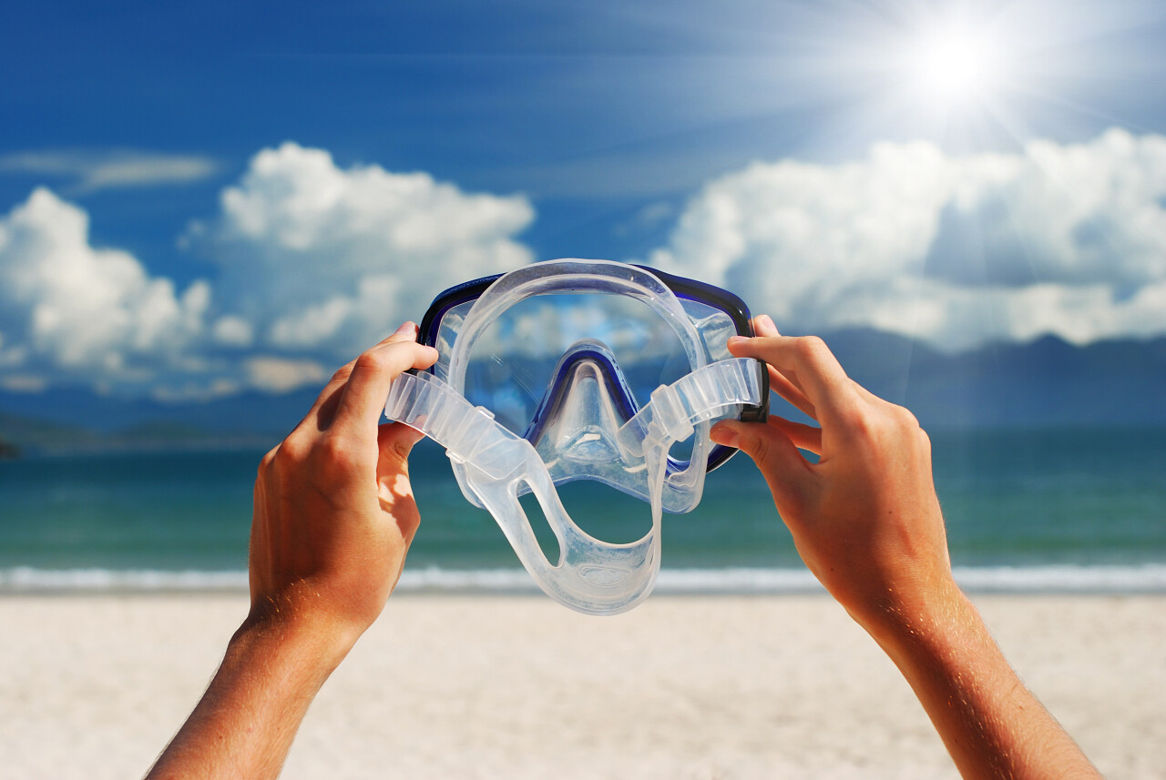 阳光下在海滩伸向天空的拿潜泳设备护目镜的手