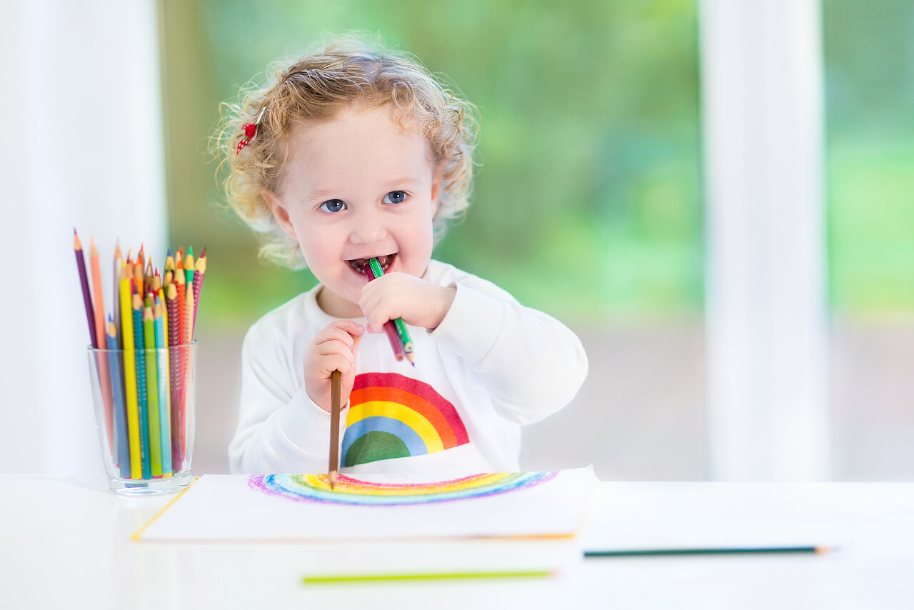 小女孩微笑着用彩色铅笔在白色桌子前画一道彩虹