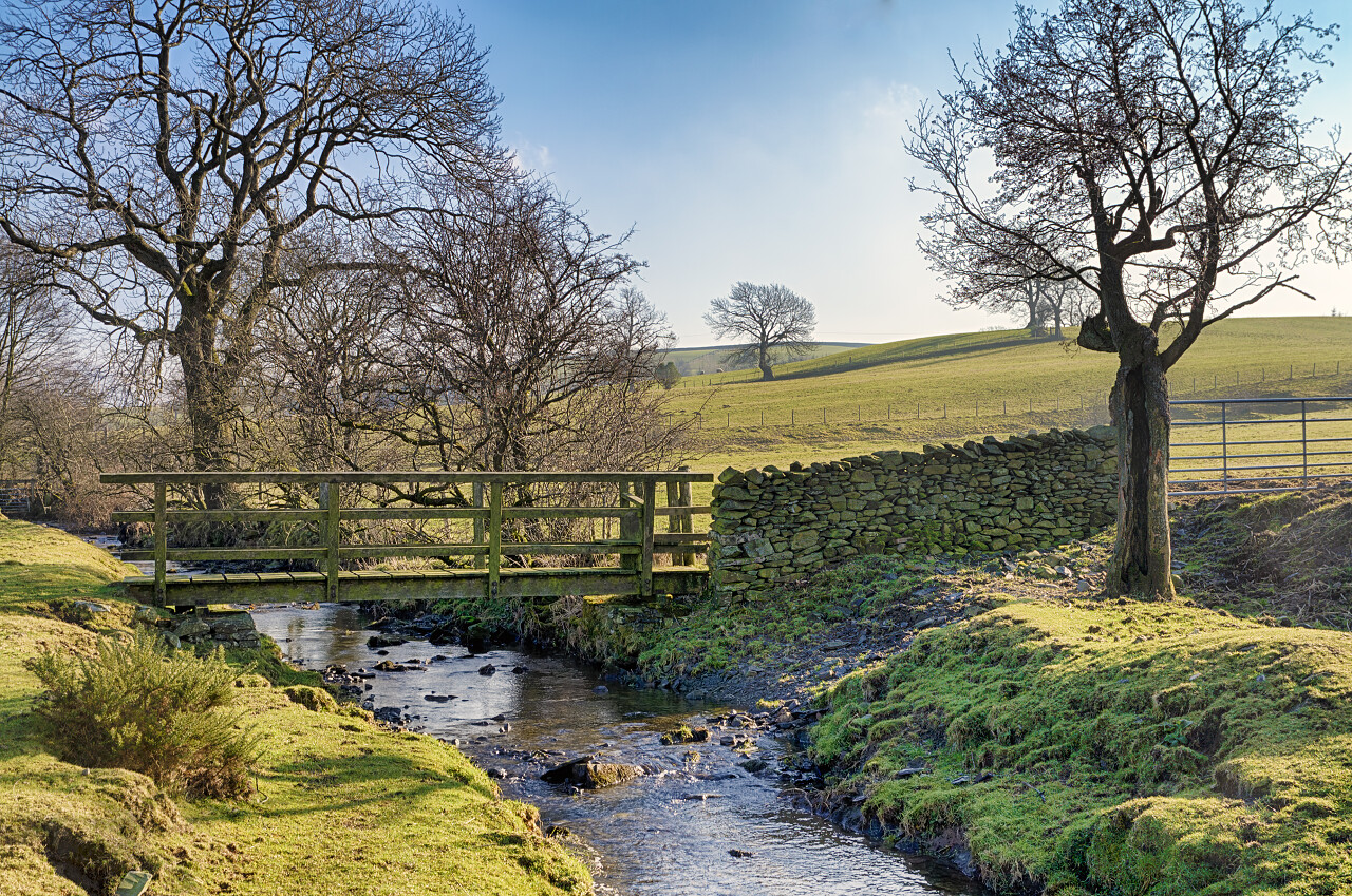 英格兰北部坎布里亚一座木制人行天桥穿过一条小溪周围环绕着乡村