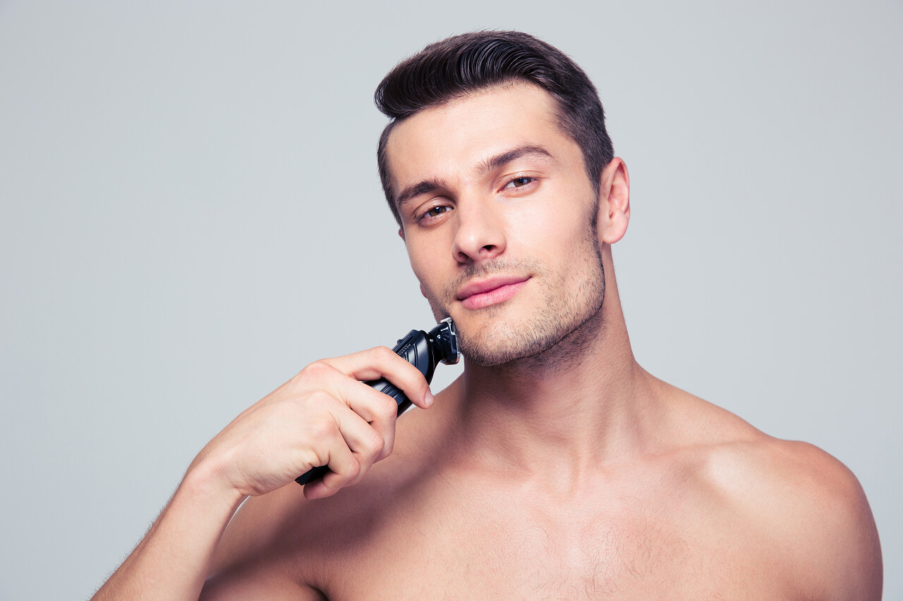 一名英俊的年轻男子用电动剃须刀一边剃须一边看着相机