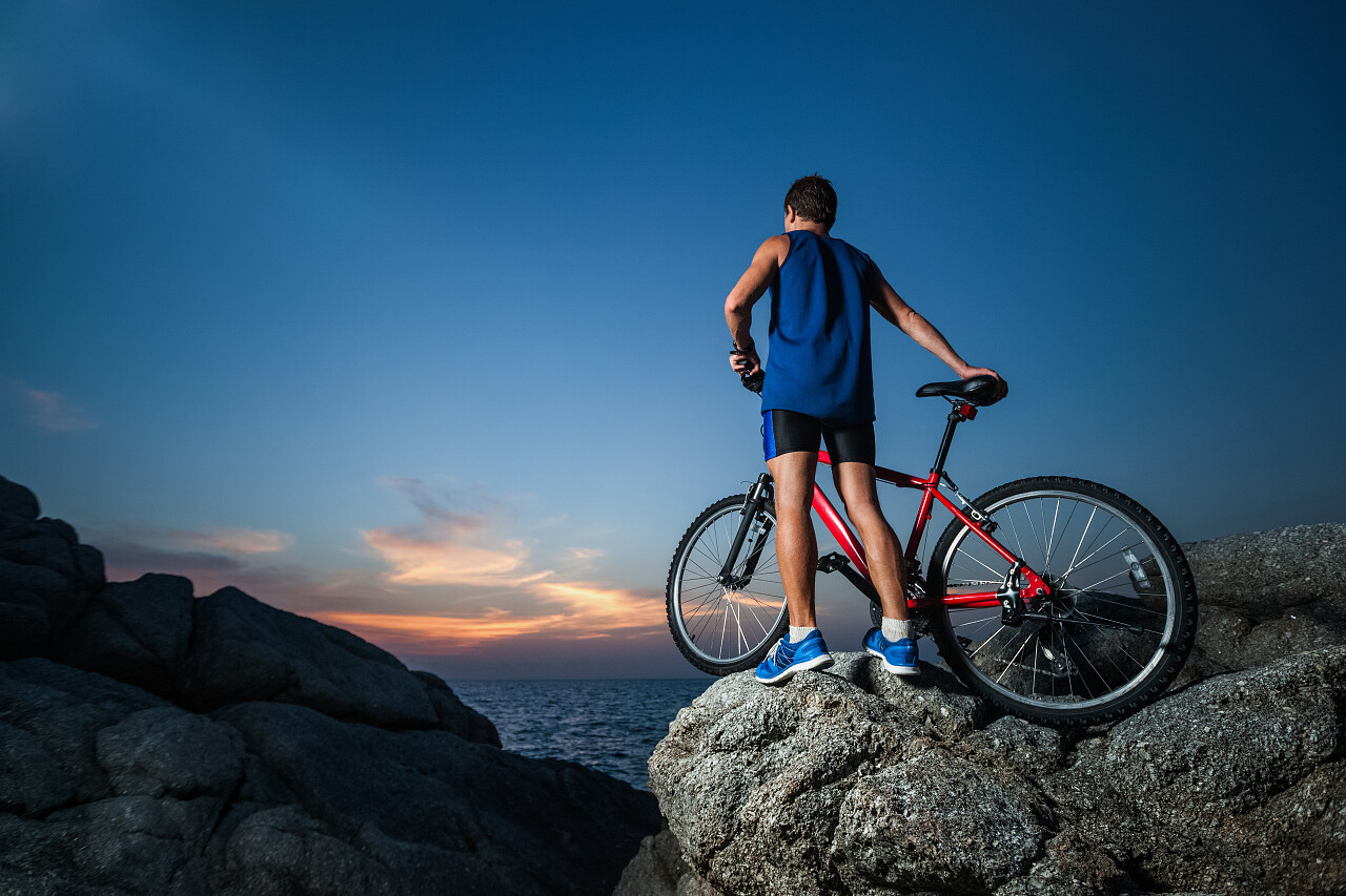 运动的人站在岩石上和一辆自行车在日落期间