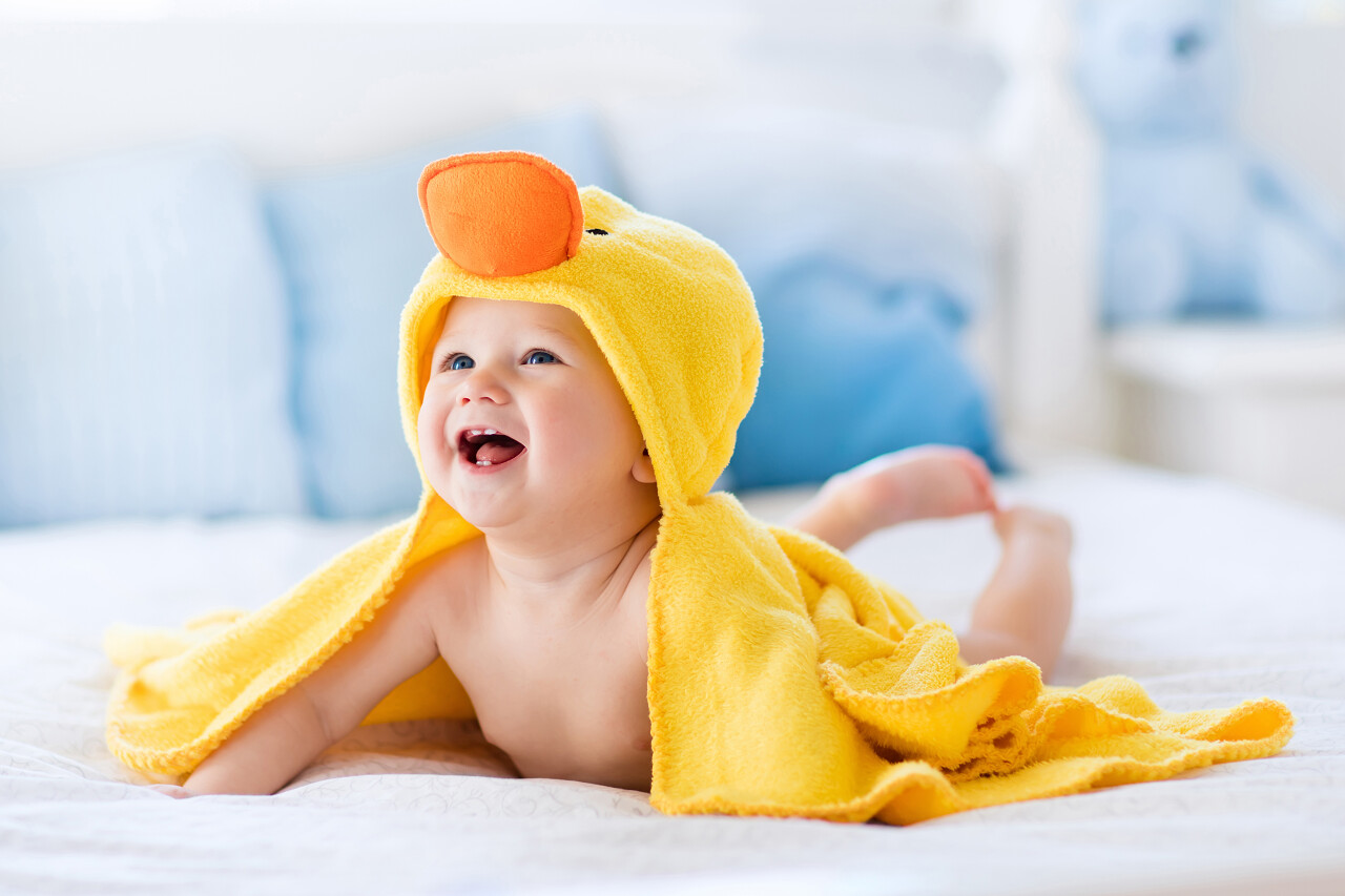 快乐的笑婴儿头戴黄色连帽的鸭毛巾沐浴或淋浴后坐在父母床上。在卧室的干净的干子。沐浴和洗衣服的小孩。儿童卫生。纺织婴儿.微笑的小孩笑脸笑容