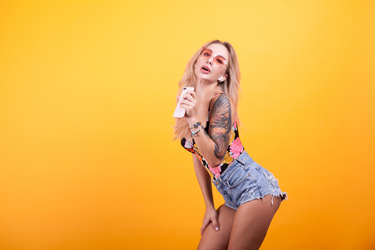 美丽可爱的女孩唱歌和听到音乐通过电话与太阳镜和帽子的黄色背景