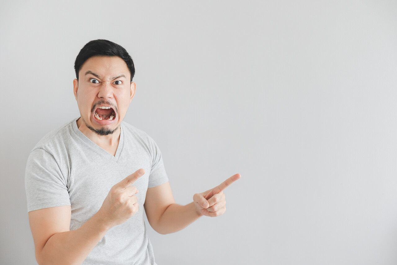 亚洲男子在灰色T恤与手点在空旷空间的愤怒的脸.