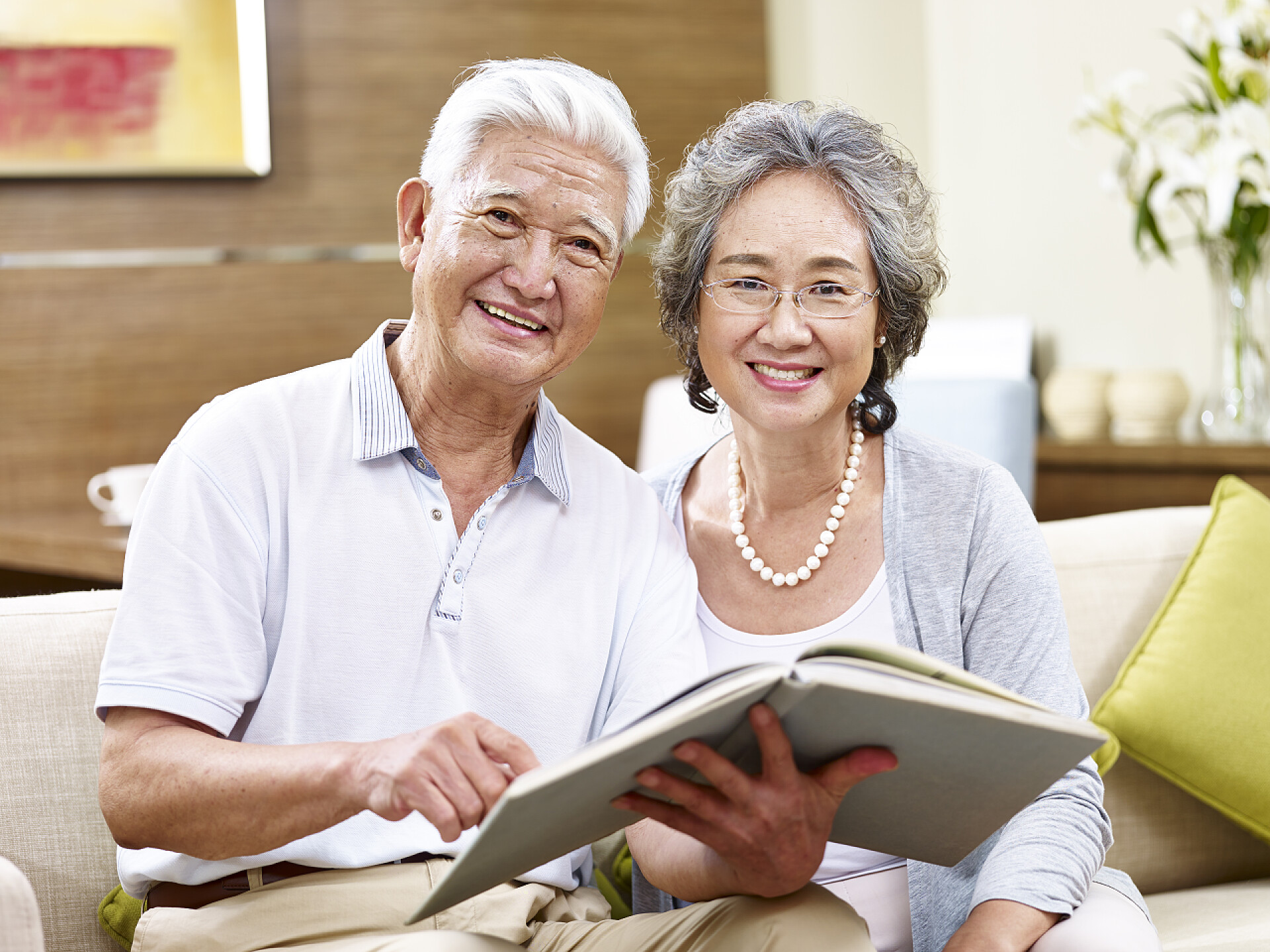亚洲老年夫妻坐在沙发上拿着书看着镜头微笑高级亚洲夫妇一起阅读一本书