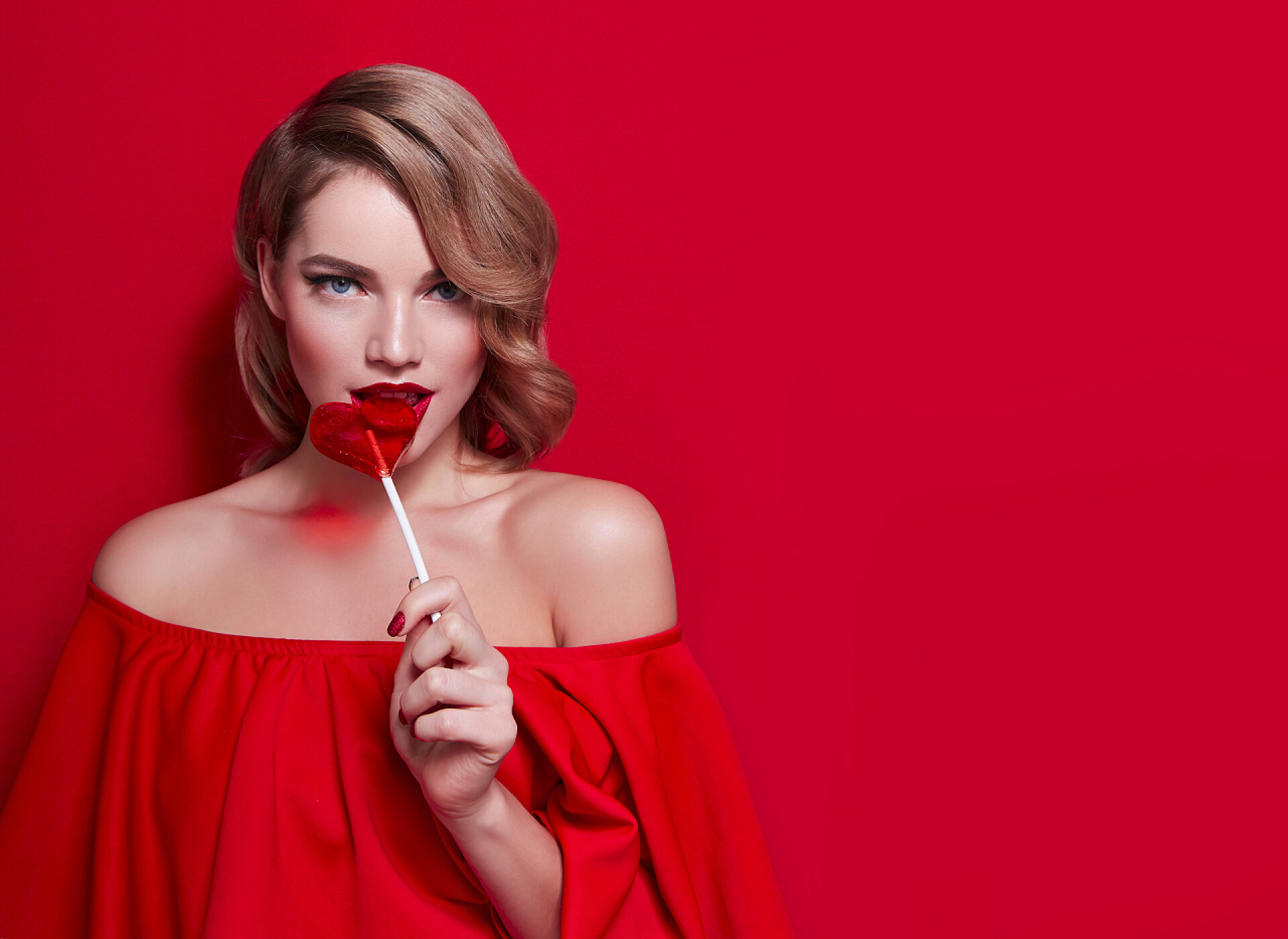 红色背景穿红色衣服的女人手里拿着一支红色心形棒棒糖女孩穿着红色红色背景上。女孩抱着他的嘴里的棒棒糖.
