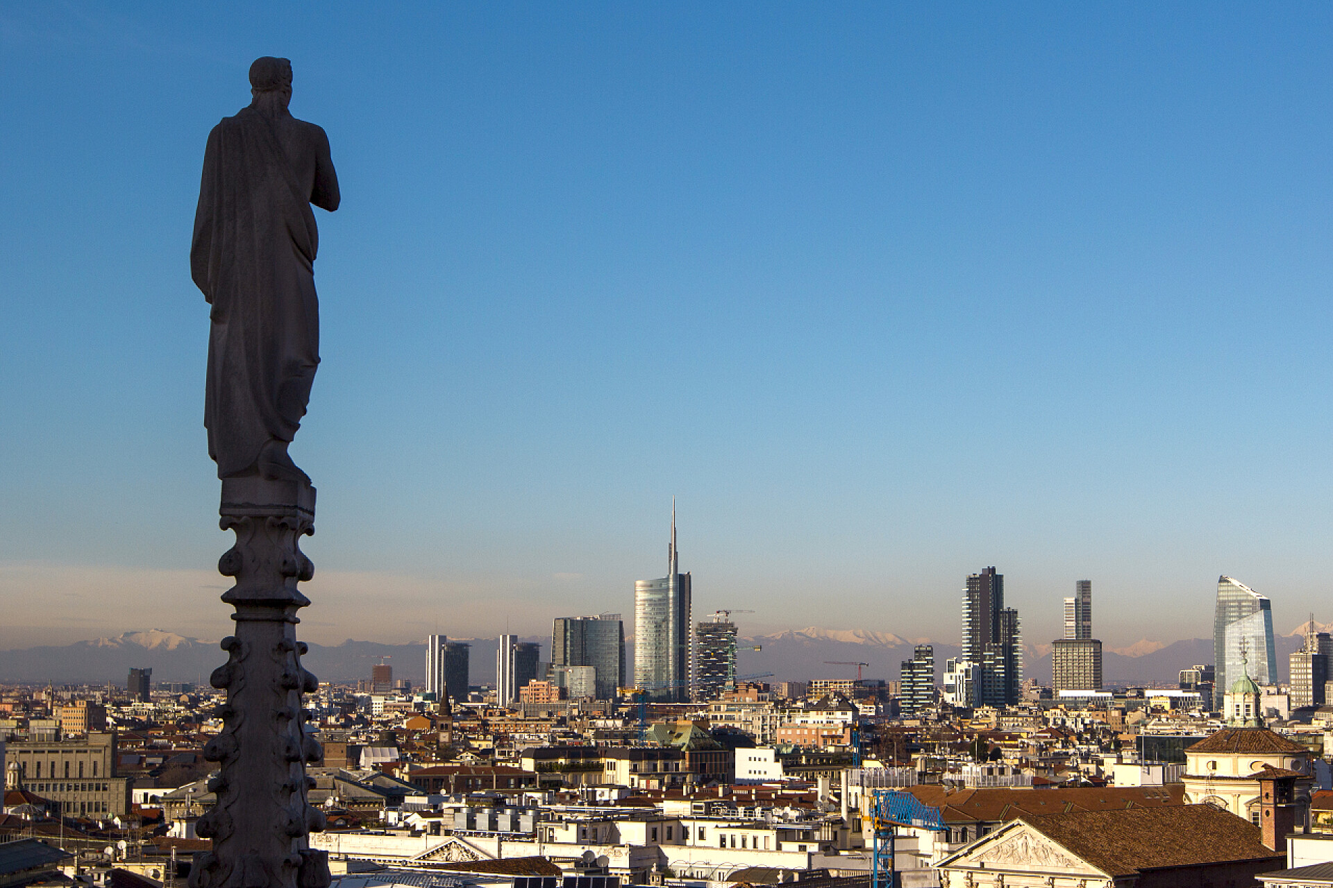 俯拍蓝天下哥特式大教堂的顶端及城市群米兰从意大利的哥特式大教堂的顶端。教堂的屋顶雕像在前景, 城市的摩天大楼在背景.