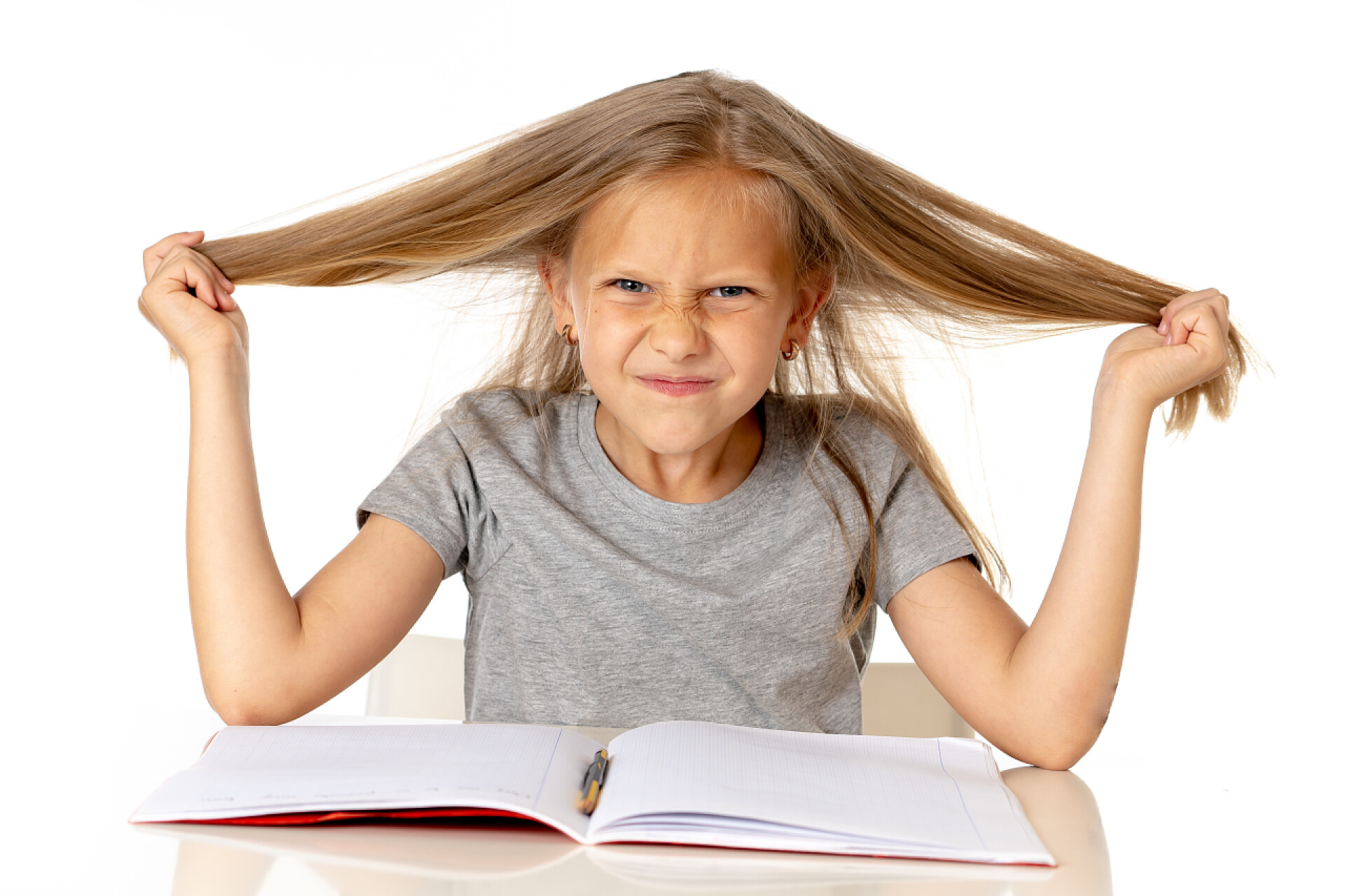白色背景前拉着头发的小女孩甜美年轻的小女生拉着她的头发绝望的压力, 而坐在学校的办公桌上做家庭作业累了, 筋疲力尽在教育理念的白色背景下孤立