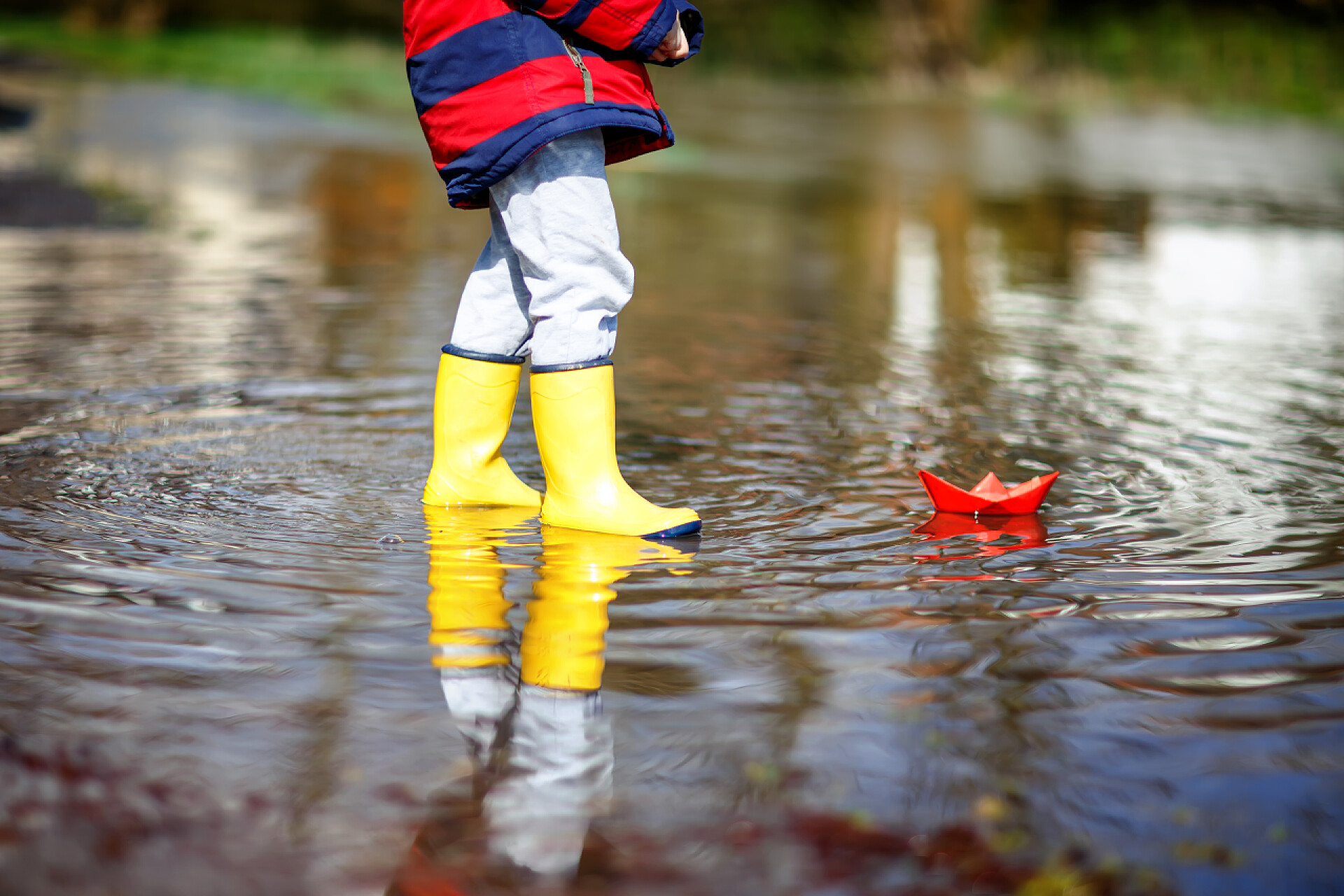 下雨的一天小男孩在户外穿着雨靴玩耍春天或秋天的一天，穿着黄色雨靴的快乐小男孩在一个巨大的水坑旁玩纸船。为孩子们提供积极的闲暇。有趣的孩子在户外玩，穿着五颜六色的衣服.