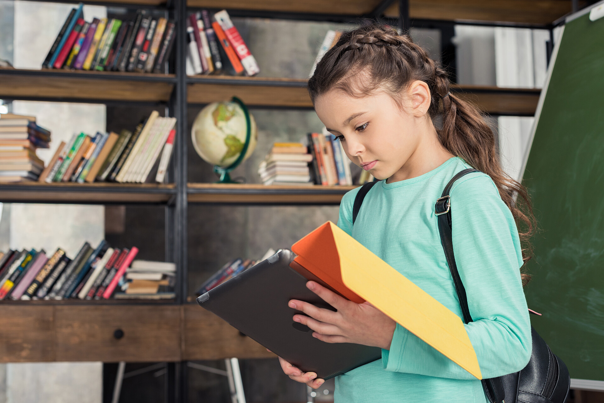 小女孩背着书包正在书架前使用平板电脑女生的数字平板电脑 