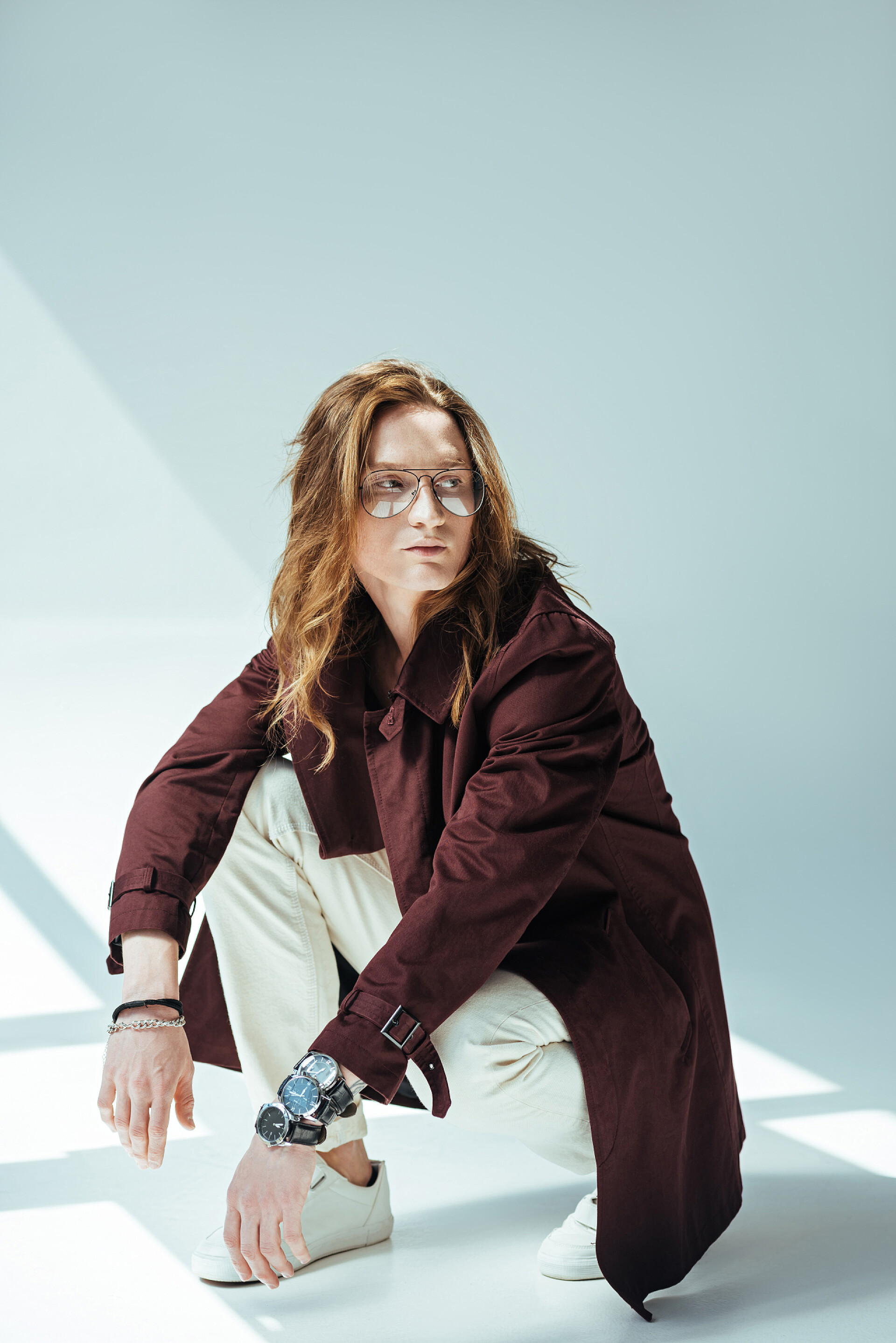 白色背景下的一个男人蹲在地上拍照时尚的人在眼镜摆在战壕大衣, 灰色