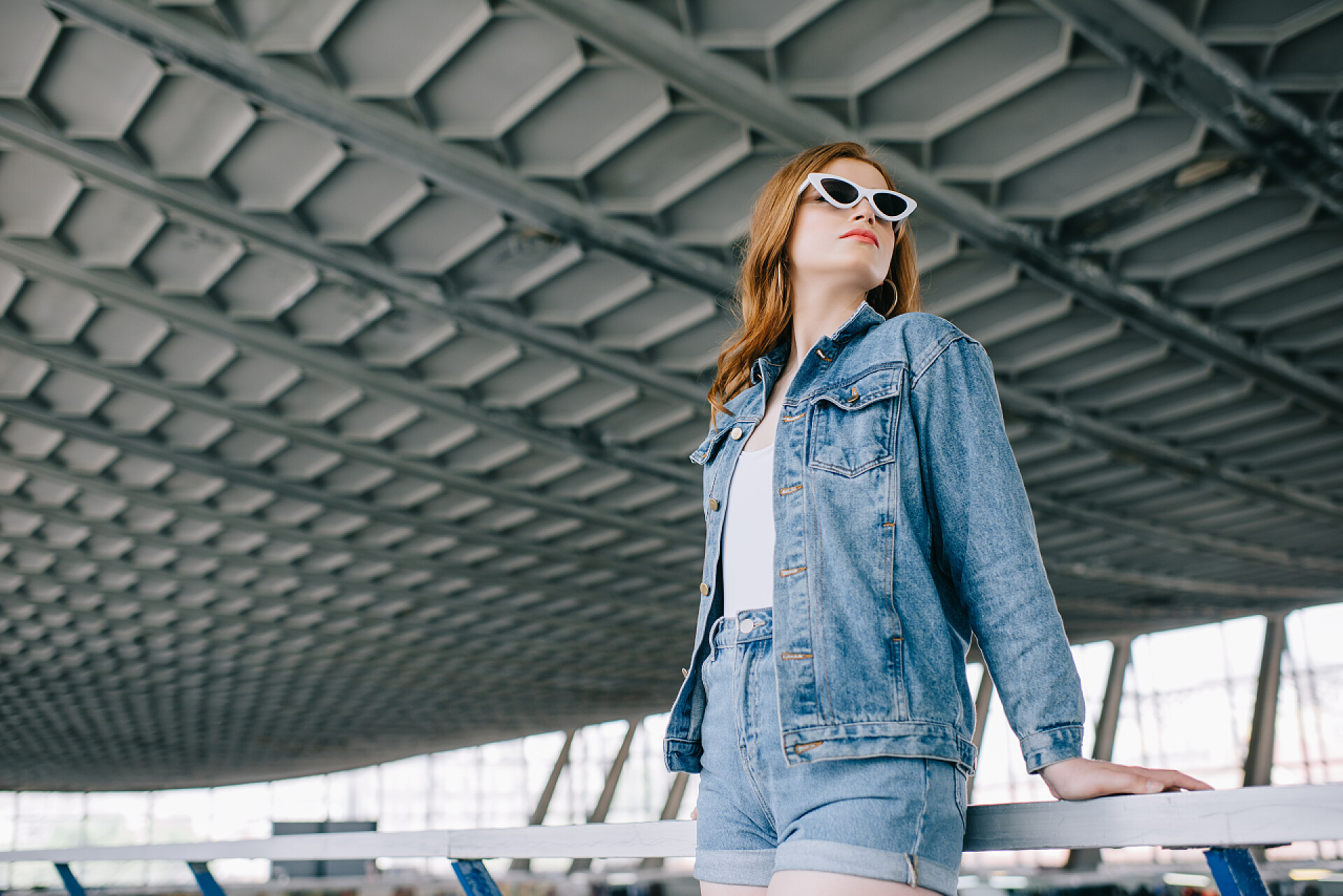 穿着牛仔服的女人站在栏杆旁牛仔服装和复古太阳镜中年轻时尚女性的低角度观