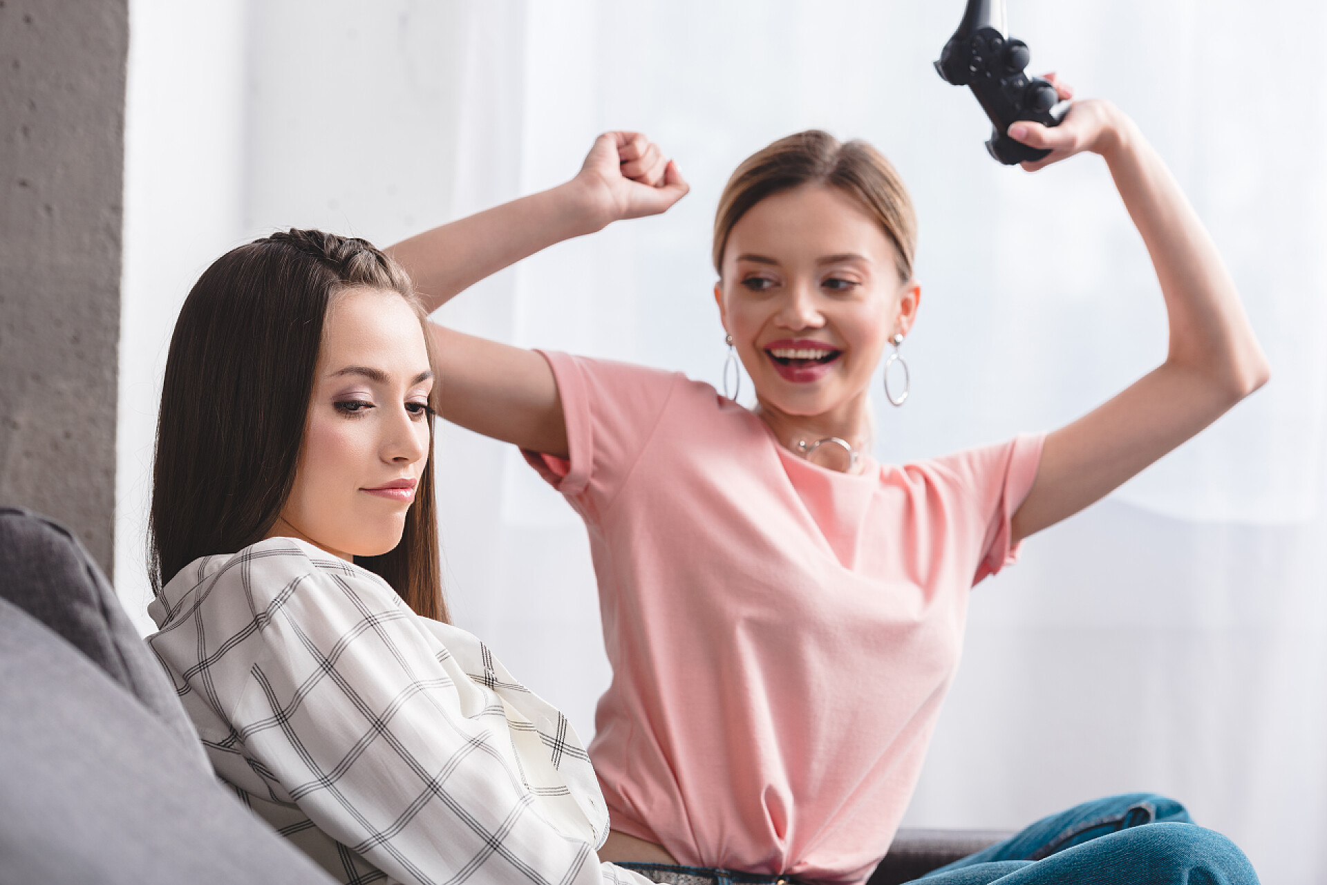 在室内打游戏的两个人微笑迷人的女人与游戏杆庆祝胜利的视频游戏和她心烦的女朋友坐在附近的家