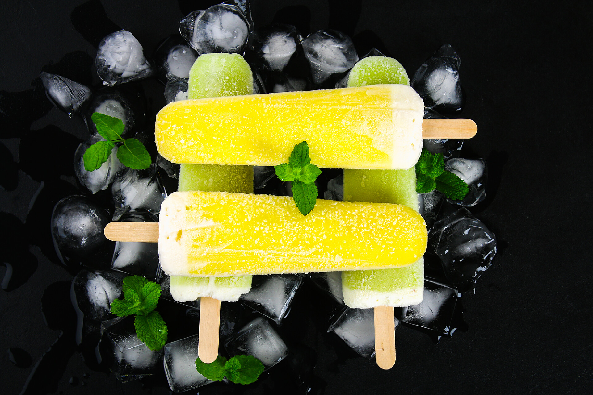 黑色桌子上的四根冰棍和冰块黄色和绿色水果冰淇淋在一根棍子与薄荷在冰在黑桌上