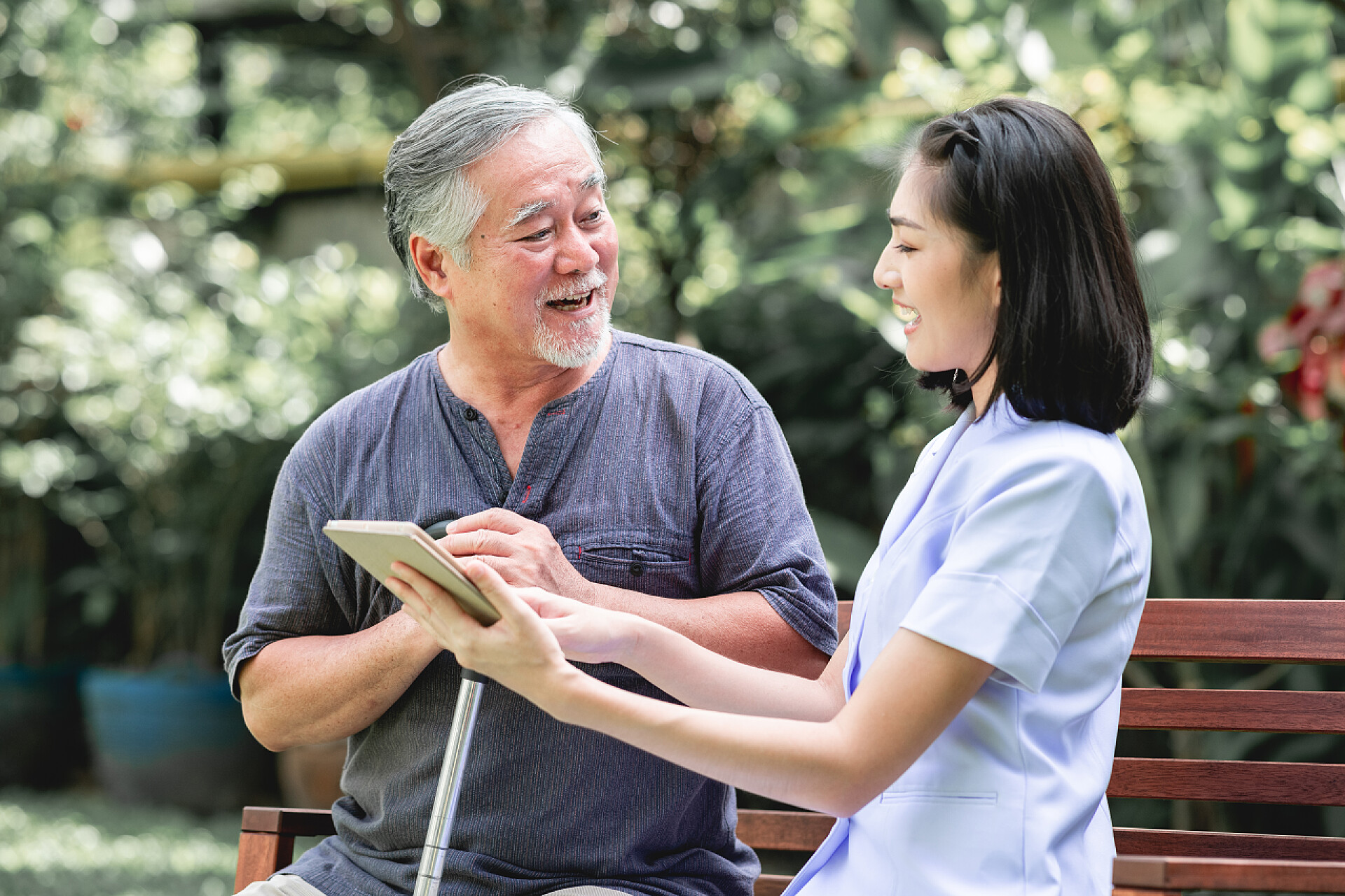 在凳子上坐着的两个人护士与病人坐在板凳上一起看片剂。亚裔老人和年轻女子坐在一起说话。放松心情.
