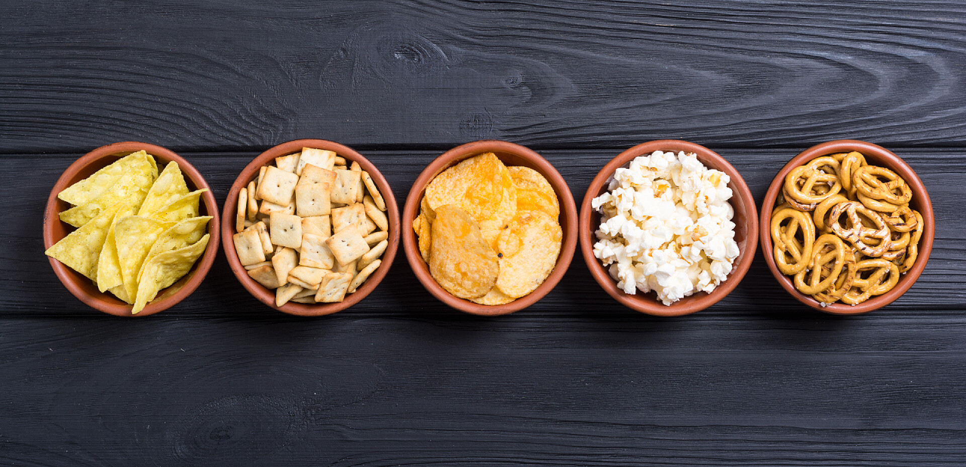 五碗在黑色桌子上的零食框架。混合零食: 椒盐脆饼、脆饼、薯片和流行玉米 