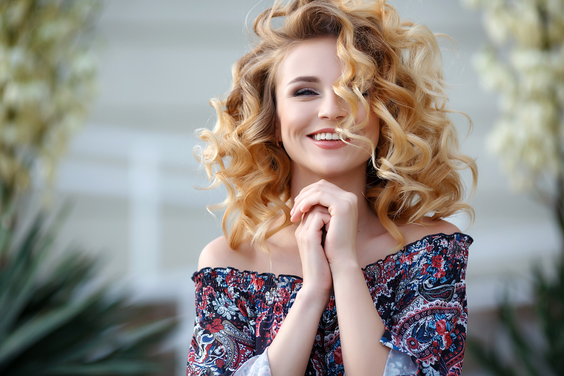 在白色背景下一个金发碧眼的美女美丽的年轻女子, 长卷发, 金发碧眼, 长长的黑色睫毛和浅色的化妆, 可爱的笑容, 穿着一件黑色的衬衫与五颜六色的图案, 开放的肩膀, 在夏天的时候在城市街道上摆姿势户外