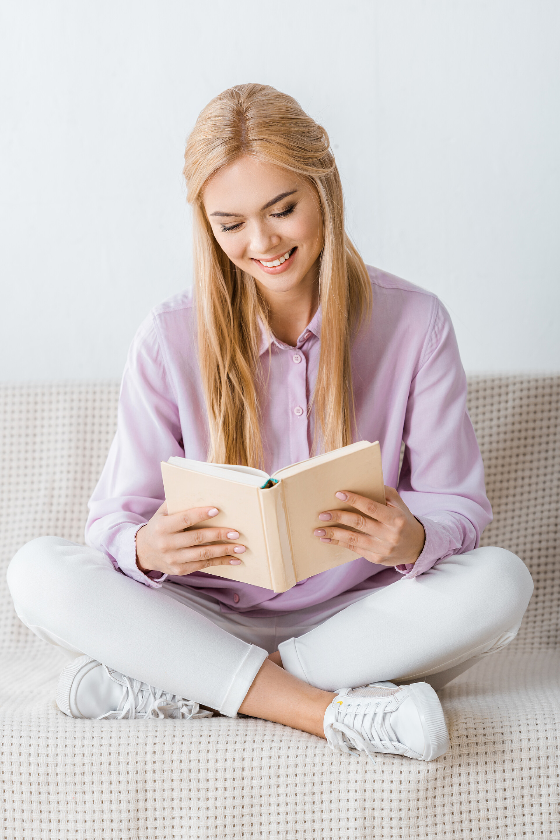 在白色背景墙前的沙发上坐着一个美丽的女人在读书美丽的微笑的女人看书, 坐在沙发上