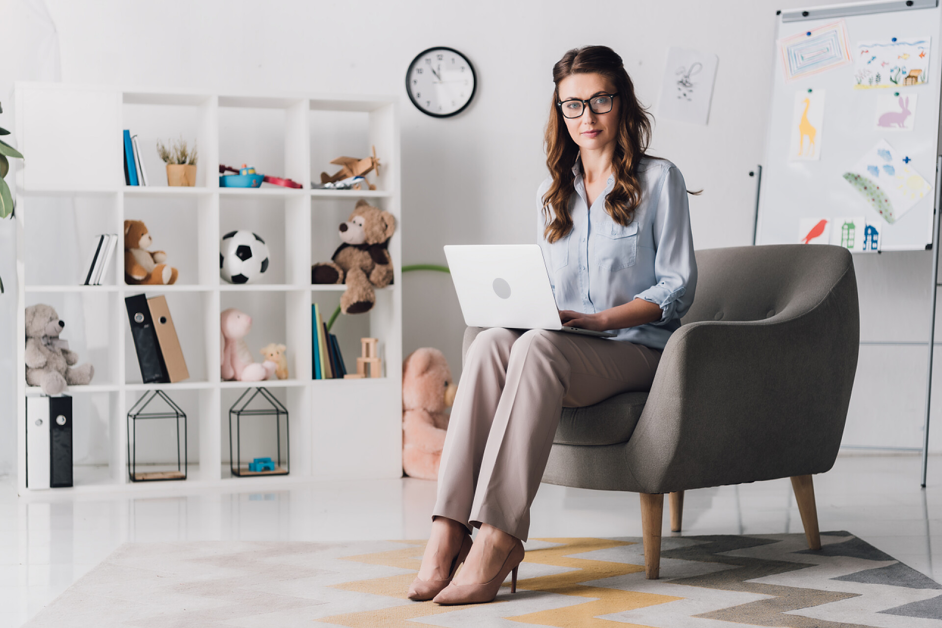 在小孩子装饰的屋子里坐着一个心理咨询师儿童心理学家与笔记本电脑的工作, 而坐在办公室的椅子上, 看着相机