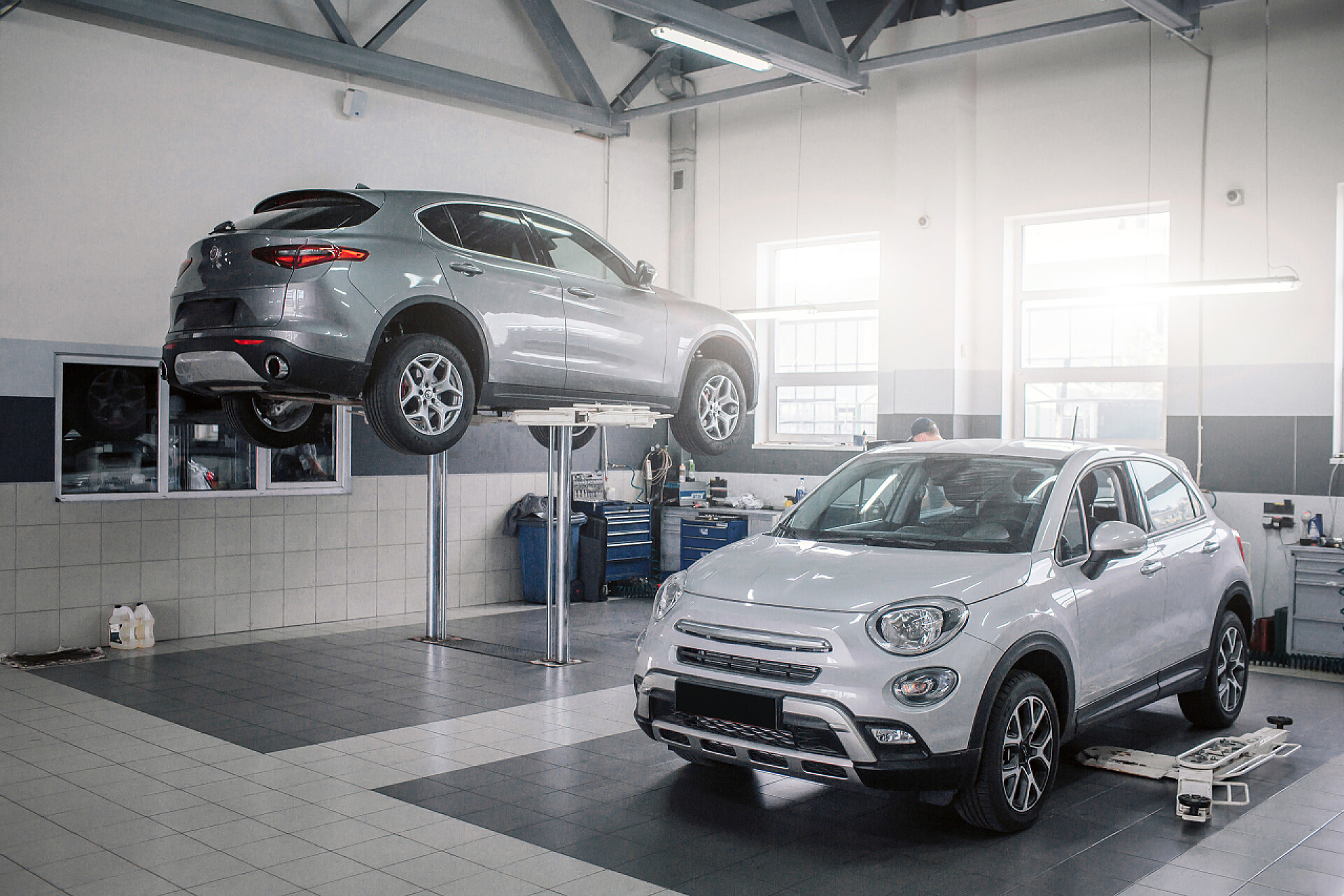 在汽车维修厂的两辆汽车白色和灰色的汽车正在建造中。他们中的一个站在站台上。戴帽子的人在白色汽车后面工作。他很专注.