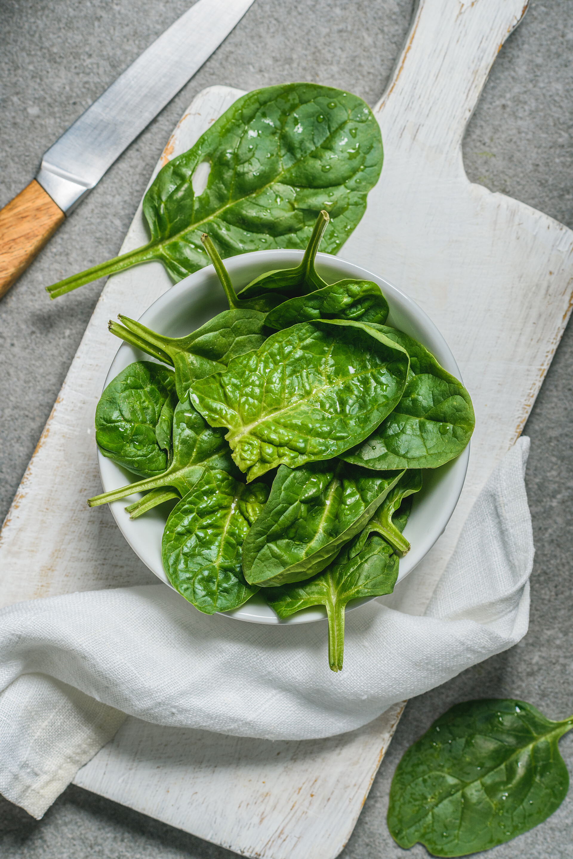 在灰色的桌子上一个木制的餐板上的蔬菜叶金属刀在白色切菜板上的碗中有机菠菜叶的顶视图