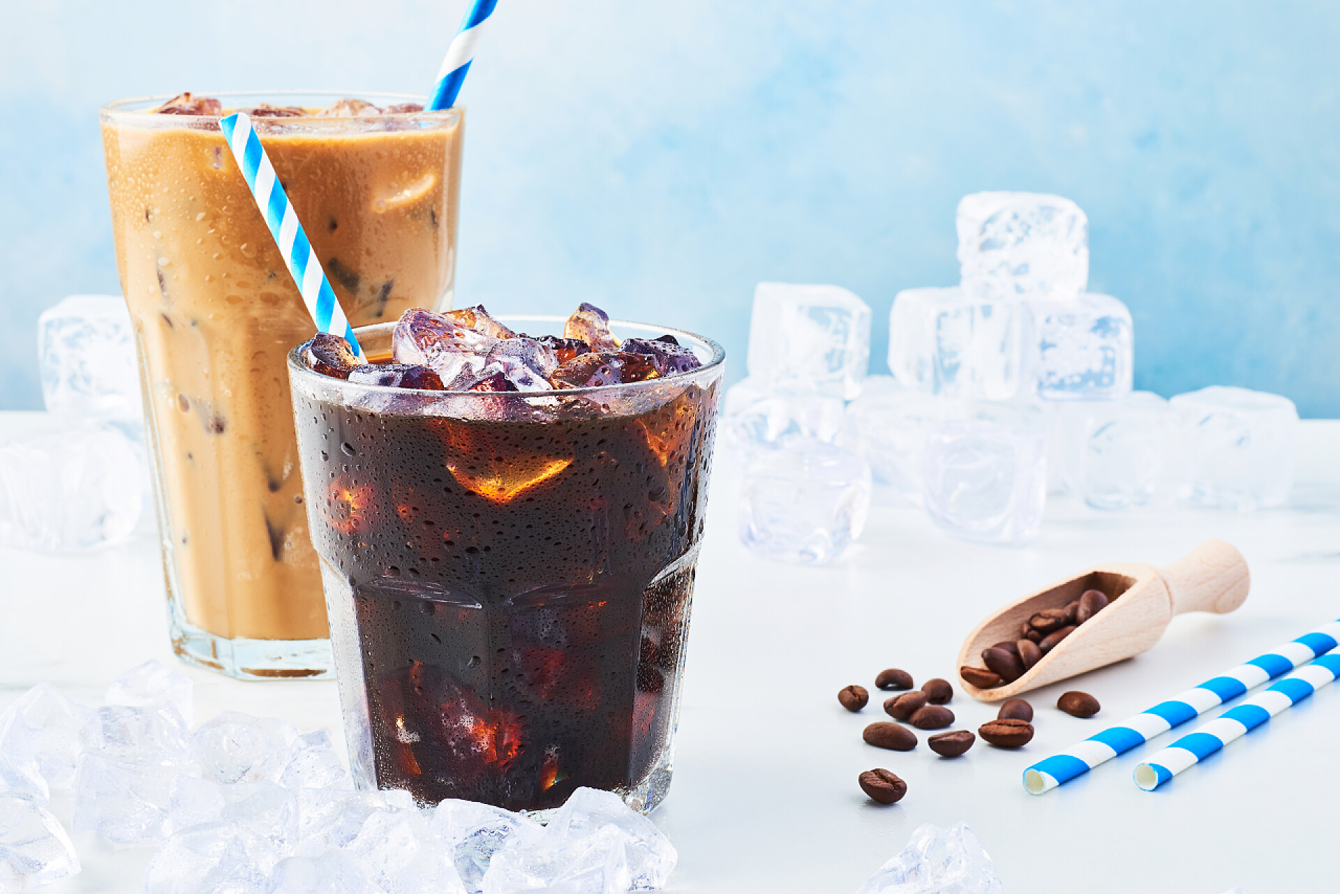 白色桌子上的两杯咖啡和冰块夏季在玻璃中喝冰咖啡, 在一个高大的杯子里喝奶油, 喝咖啡豆和用吸管在蓝色背景的白色大理石桌子上围住冰。复制文本的空间.