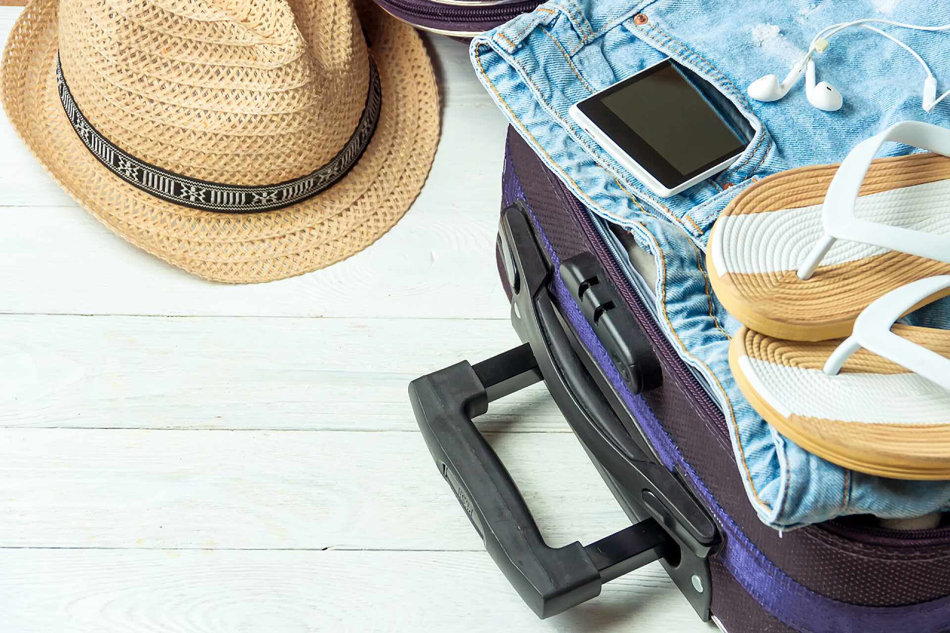 在白色的地板上有着一个行李箱上面有牛仔裤拖鞋手机旁边有帽子打开行李箱, 在白色木桌上配有旅行配件, 并配有仿制空间.