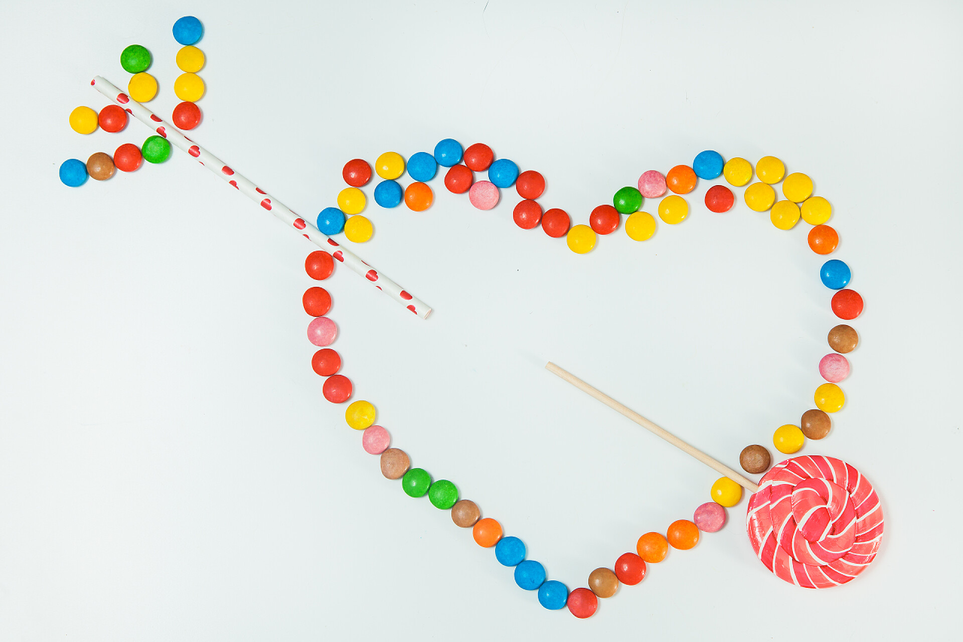 巧克力和棒棒糖组成的一个箭穿过爱心的图案白色背景平铺心箭头的巧克力糖果棒棒糖