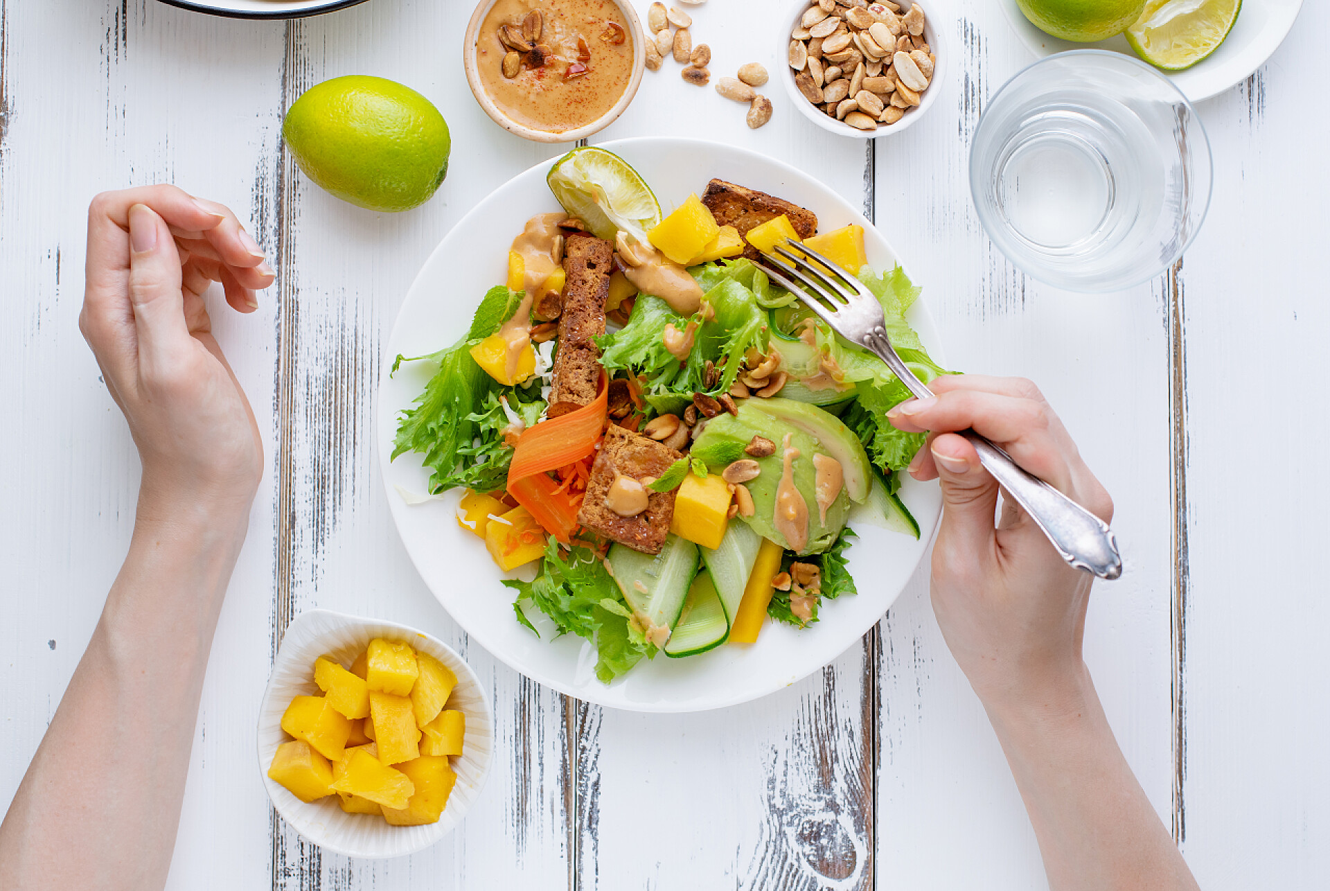 女性手拿叉子品尝美味水果沙拉女性双手拿着叉子在一盘新鲜的沙拉与芒果和鳄梨与豆腐和甜花生酱在白色的木桌上