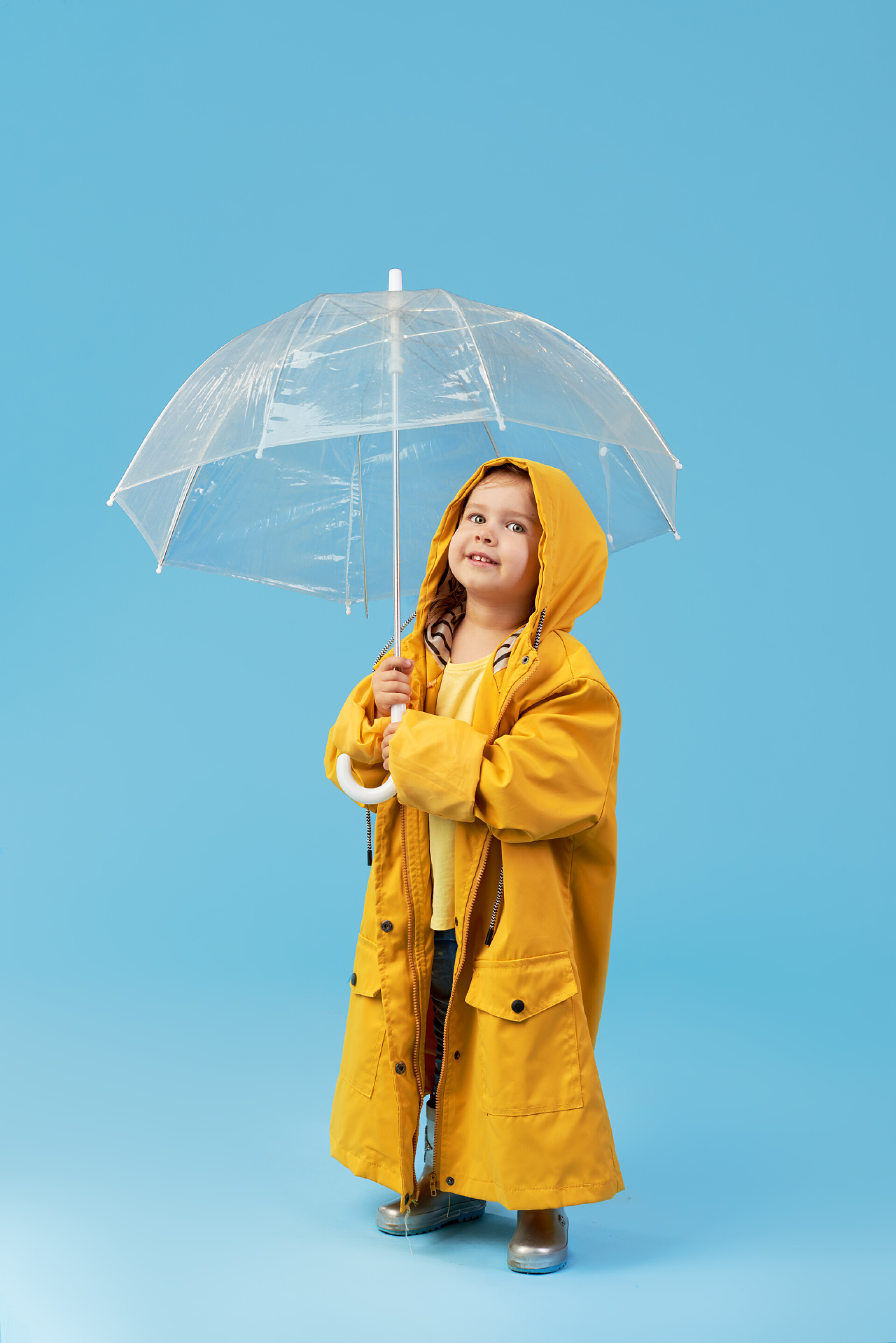 蓝色背景上的孩子打着伞快乐有趣的孩子,在蓝色的工作室背景上摆出透明的伞.女孩穿着黄色雨衣和橡胶靴。持有一个老式旅行箱
