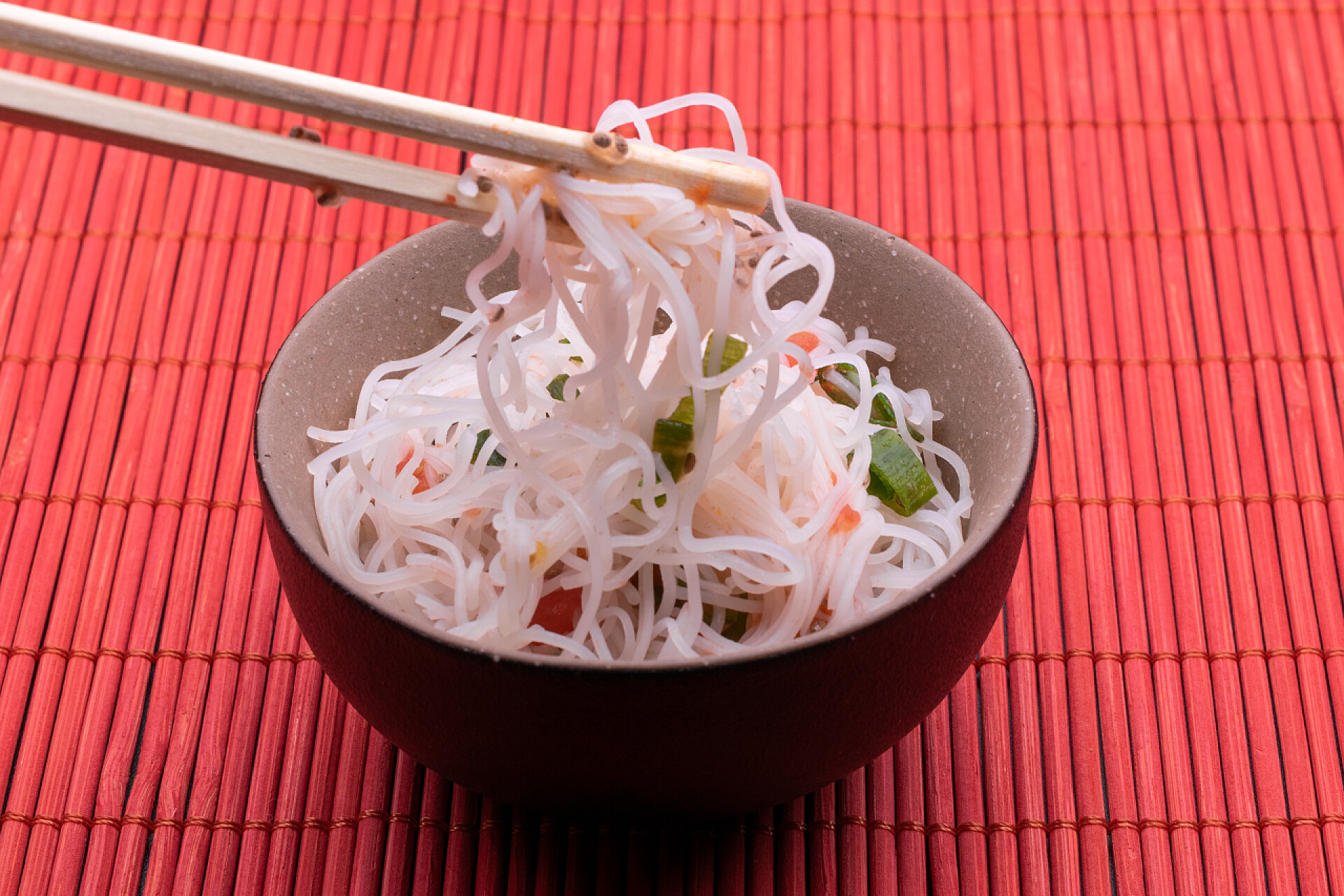 亚洲沙拉面亚洲沙拉，米饭面条和蔬菜。煮熟的米面泰式在黑色和棕色的碗与筷子隔离在红色木制背景. 