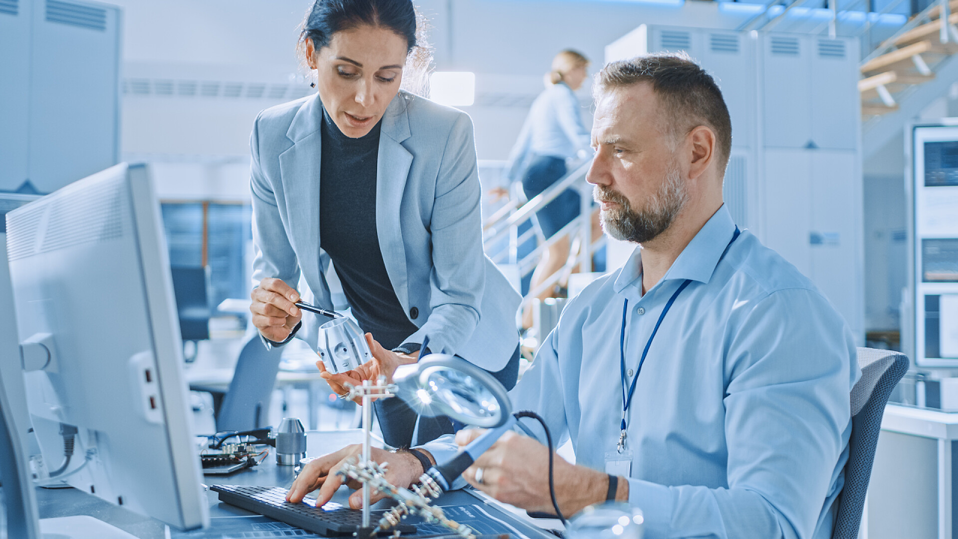 研究机器人开发的工程师女工业工程师与电子专家交谈，她持有金属部件原型，他在电脑上工作。现代明亮的办公室与时尚的人