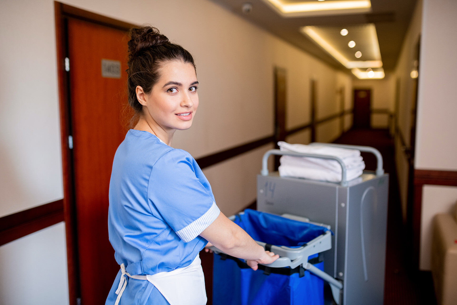 酒店走廊打扫卫生的服务员早上，穿着制服的年轻的黑发女服务员在酒店的走廊边推推车的时候看着你