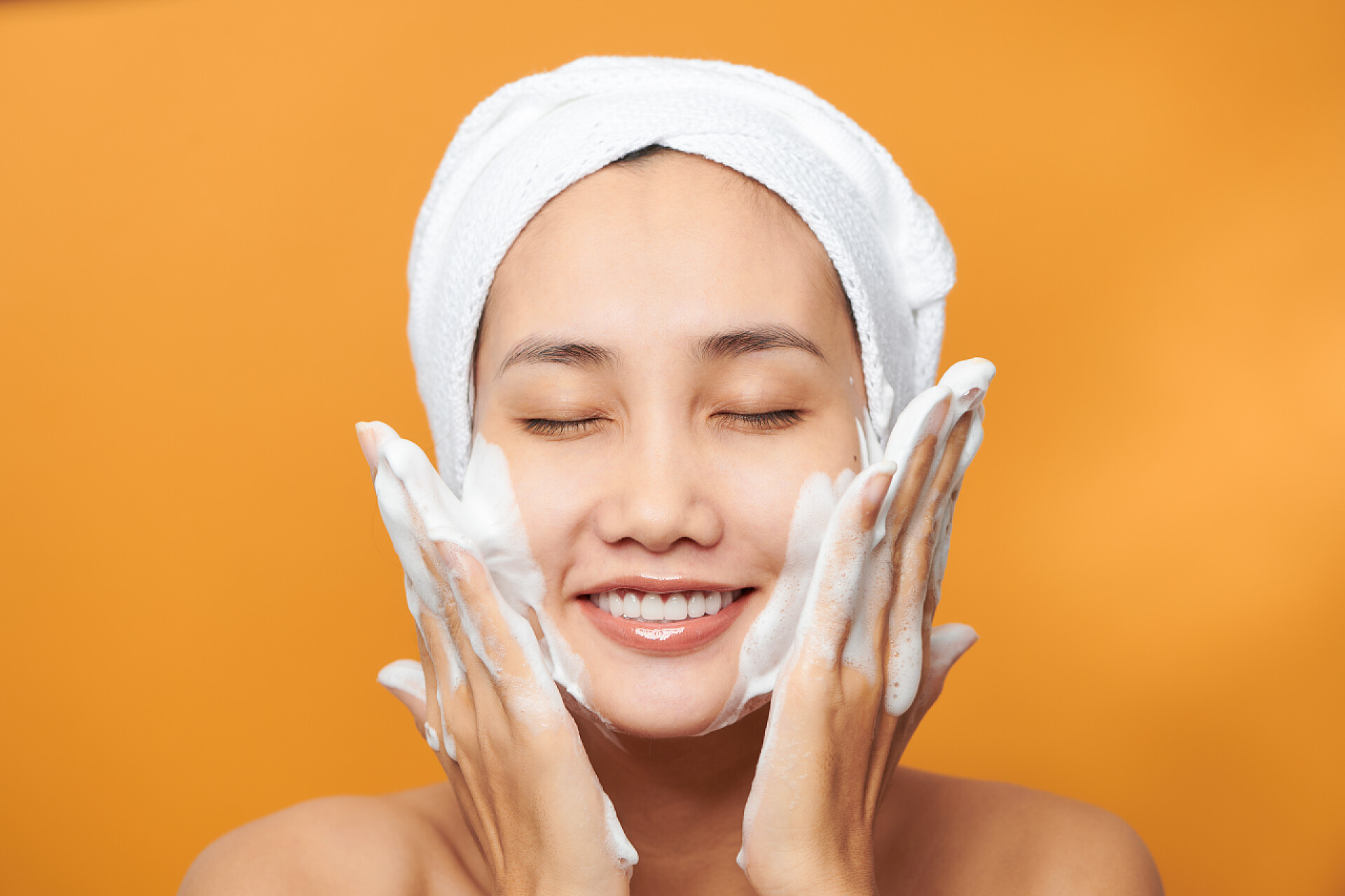 橙色背景上用洗面奶洗脸的美女Happy young Asian woman applying face cream while wearing a towel and touching her face. Isolated on orange background