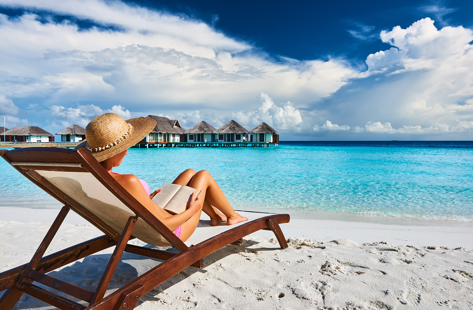 蓝天白云下在海滩边上坐在沙滩椅上j看书的美女读一本书在海滩上的年轻女子