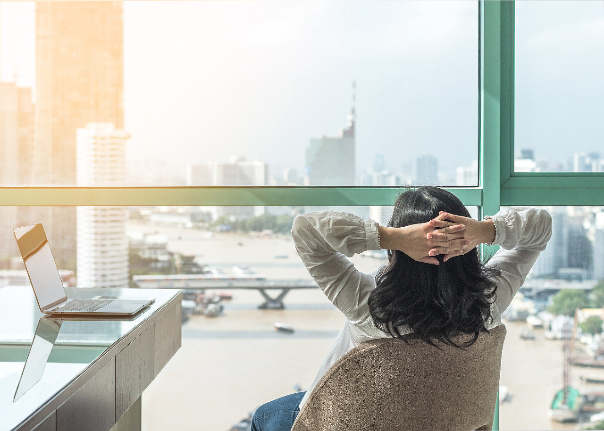 抱头看窗外的女人在家里办公室的豪华商务酒店里，女性可以轻松地工作和休息，办公室里桌上有电脑笔记本电脑