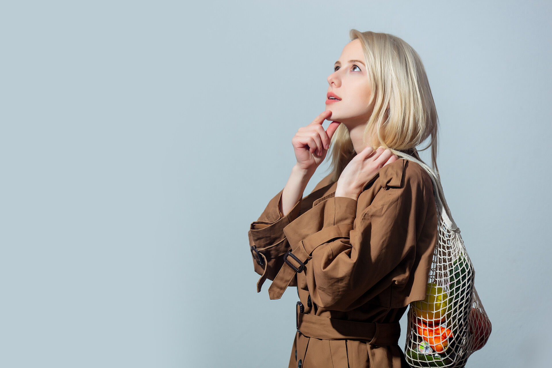 灰色背景上穿披风抬头抚摸下巴的时尚女子特写穿着斗篷的金发姑娘拿着灰色背景的水果和网袋 