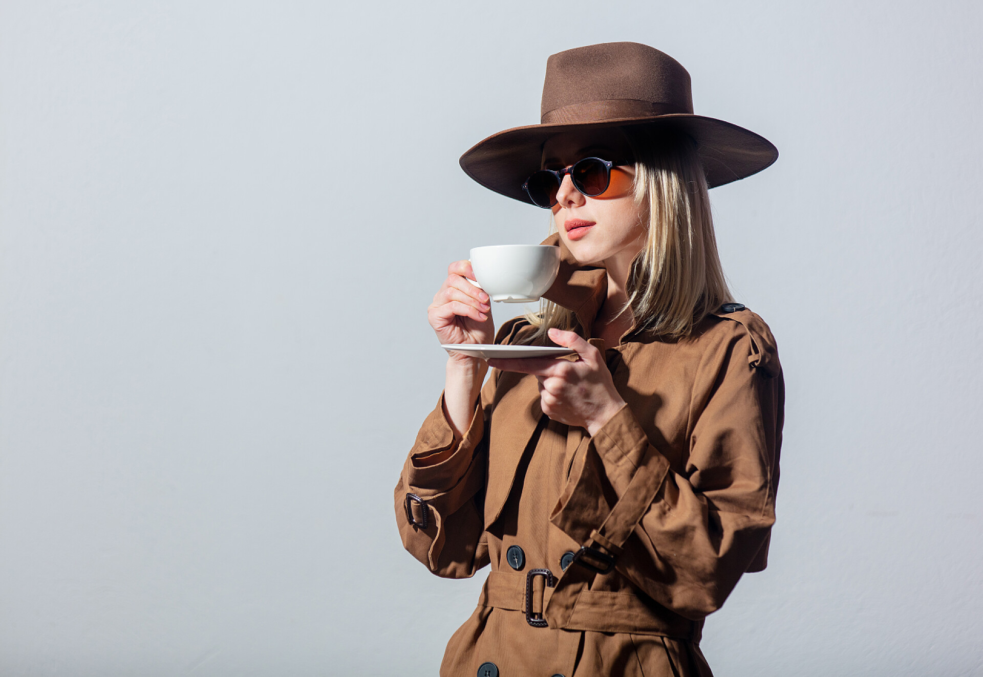 灰色背景上穿披风戴帽子喝咖啡的时尚女子特写穿着斗篷、帽子和太阳镜的时尚女人拿着一杯灰色背景的咖啡