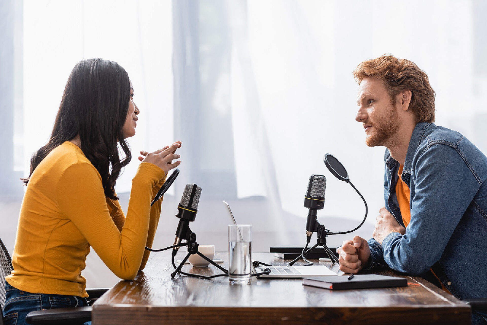 明亮的播音室内两个工作人员正在交谈亚洲年轻女子在演播室接受采访时与红头发播音员交谈的侧视图