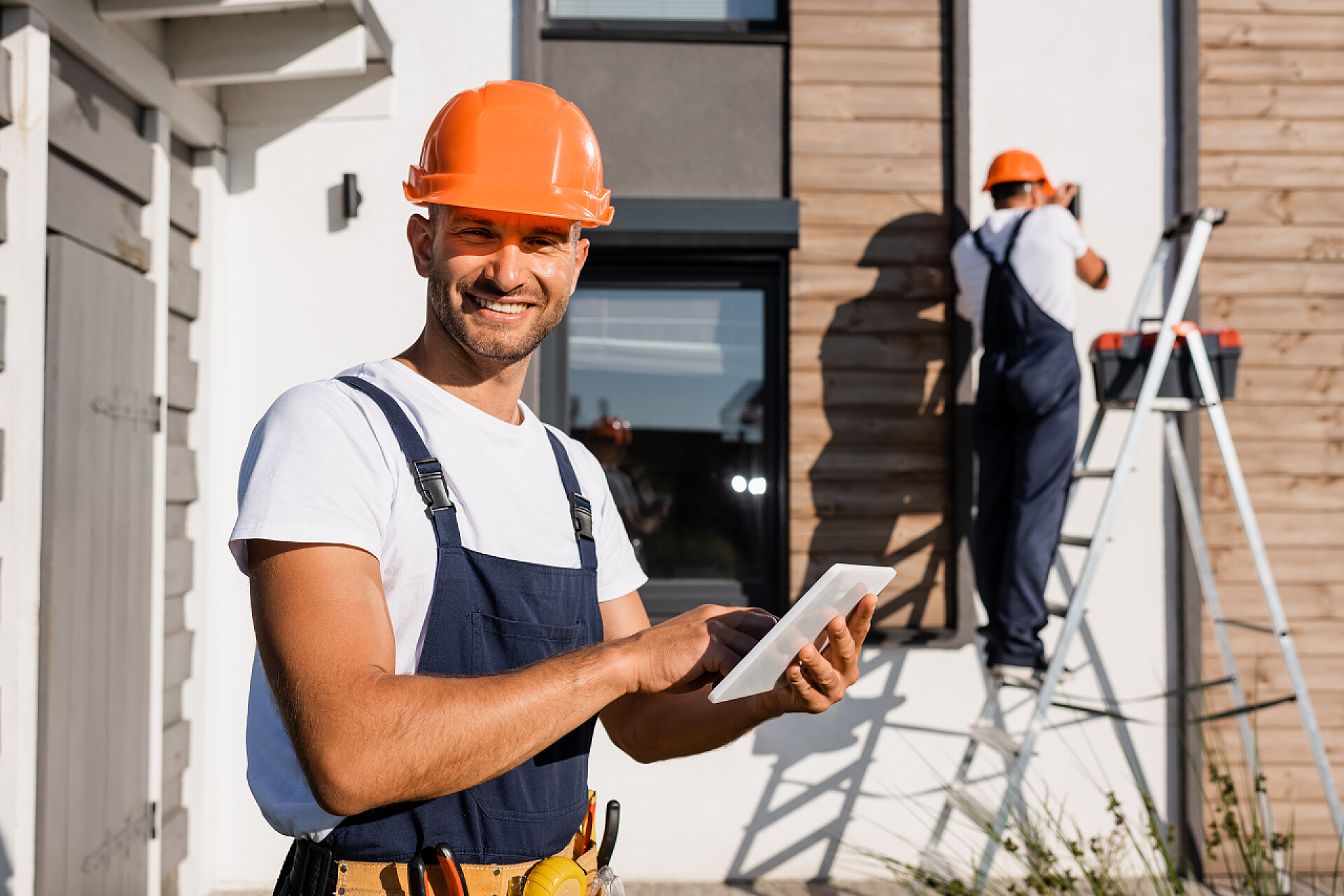 两个工人在讨论房子的建筑带数码平板电脑的建筑商的选择性聚焦，当同事在房子附近的梯子上做背景时，他们看着相机 