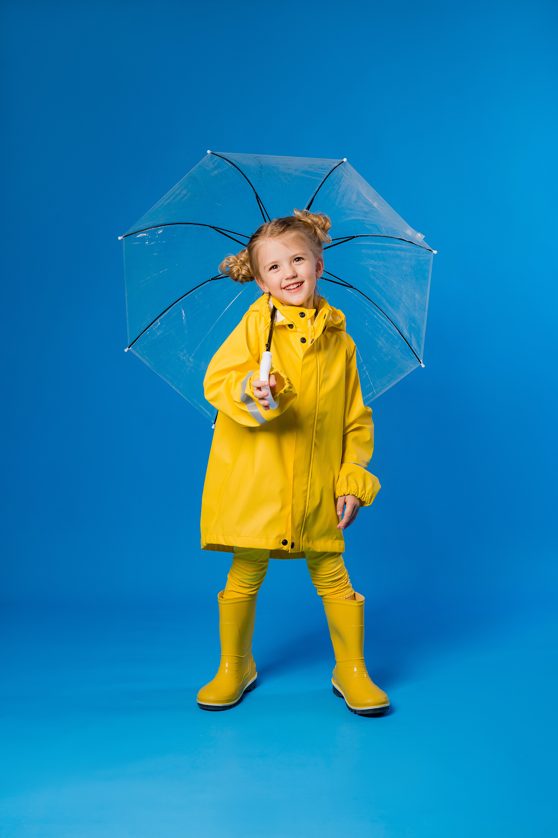 穿着黄色衣服的女孩在拍照穿着雨衣和橡胶靴的快乐女孩站在蓝色的背景上，带着一把透明的雨伞。复制空间图片并放置到您的广告文本中