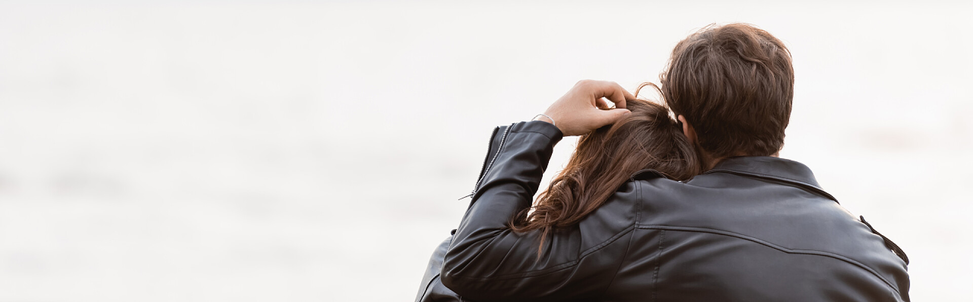 在大海边互相依靠的情侣男人在海面附近的皮夹克里摸着女朋友的头发 