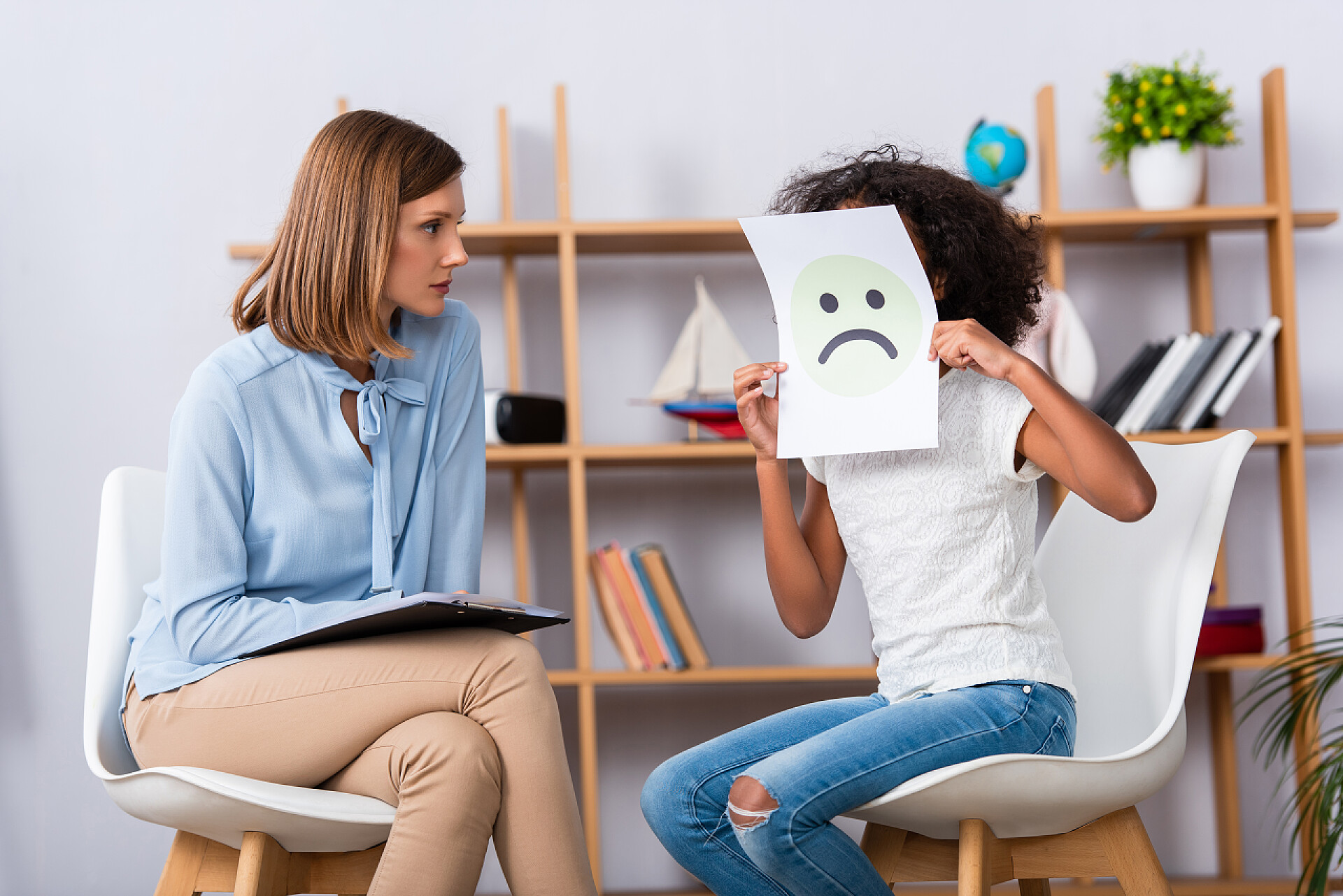 儿童保健在咨询室里的女孩拿着哭脸心理学家看着非洲裔美国女孩，她满脸愁容，背景上的办公室模糊不清