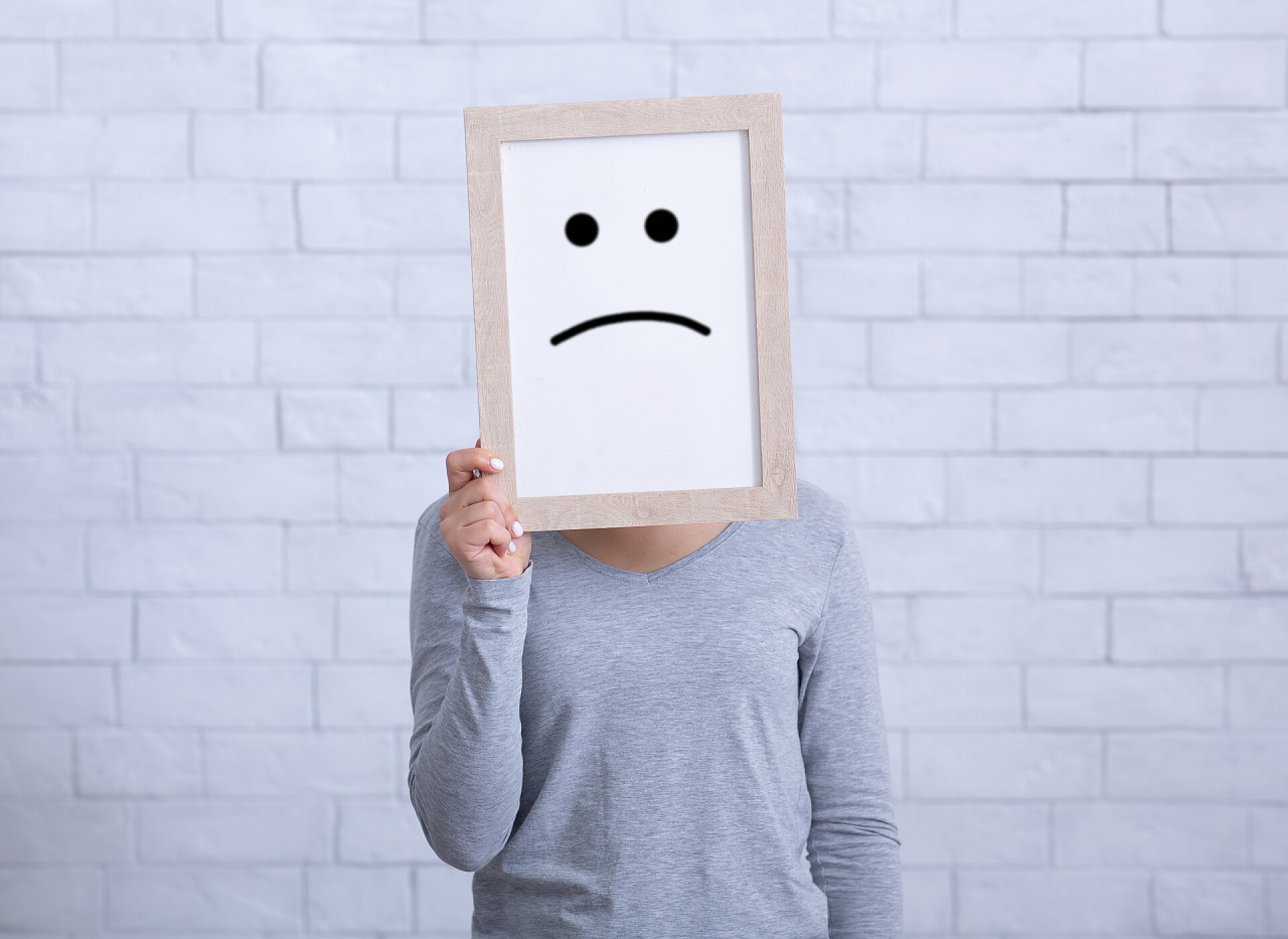 白色背景墙前的女人Millennial lady holding picture frame with unhappy emoticon, covering her face against white wall