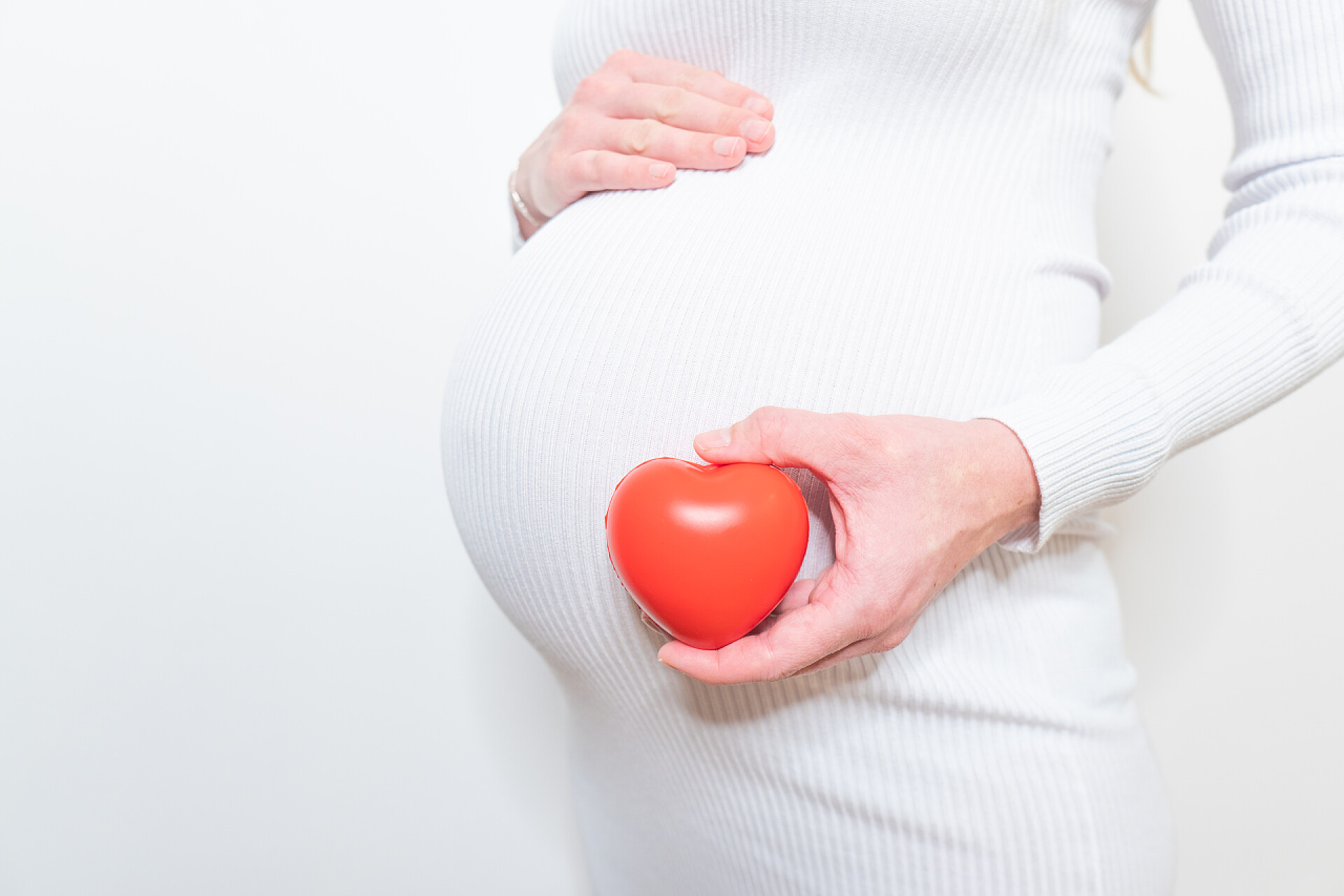 白色背景墙的一个美丽孕妇拿着红心在阳光明媚的夏日，怀孕、分娩、家庭观念、孕妇和户外心脏符号